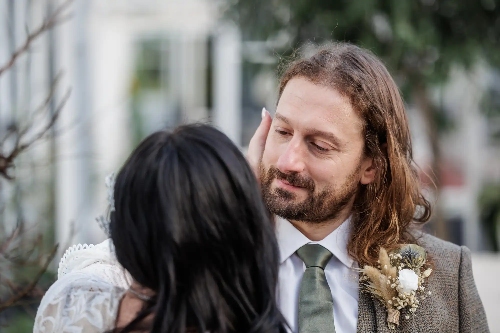 A man with long brown hair, a beard, and a suit with a buttonhole gazes lovingly at a woman with long dark hair, whose face is turned away from the camera. They appear to be outside, surrounded by greenery.