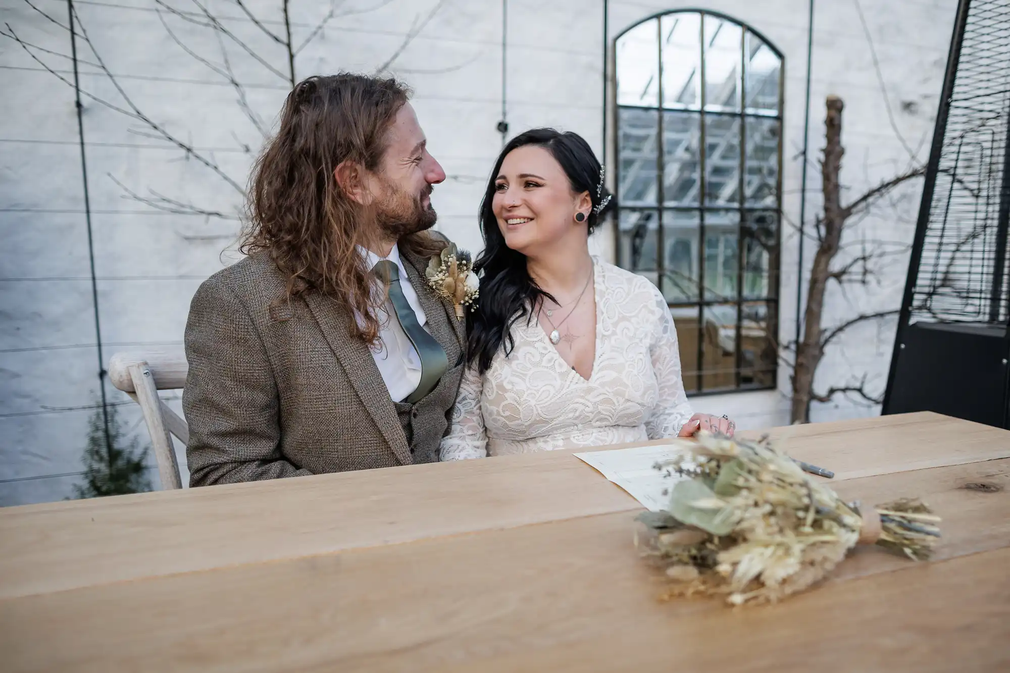 A smiling couple in wedding attire sit at a wooden table outdoors. The bride holds a pen and wears a white lace dress; the groom wears a brown suit. A dried bouquet rests on the table. They look at each other lovingly, with large windows and bare branches in the background.
