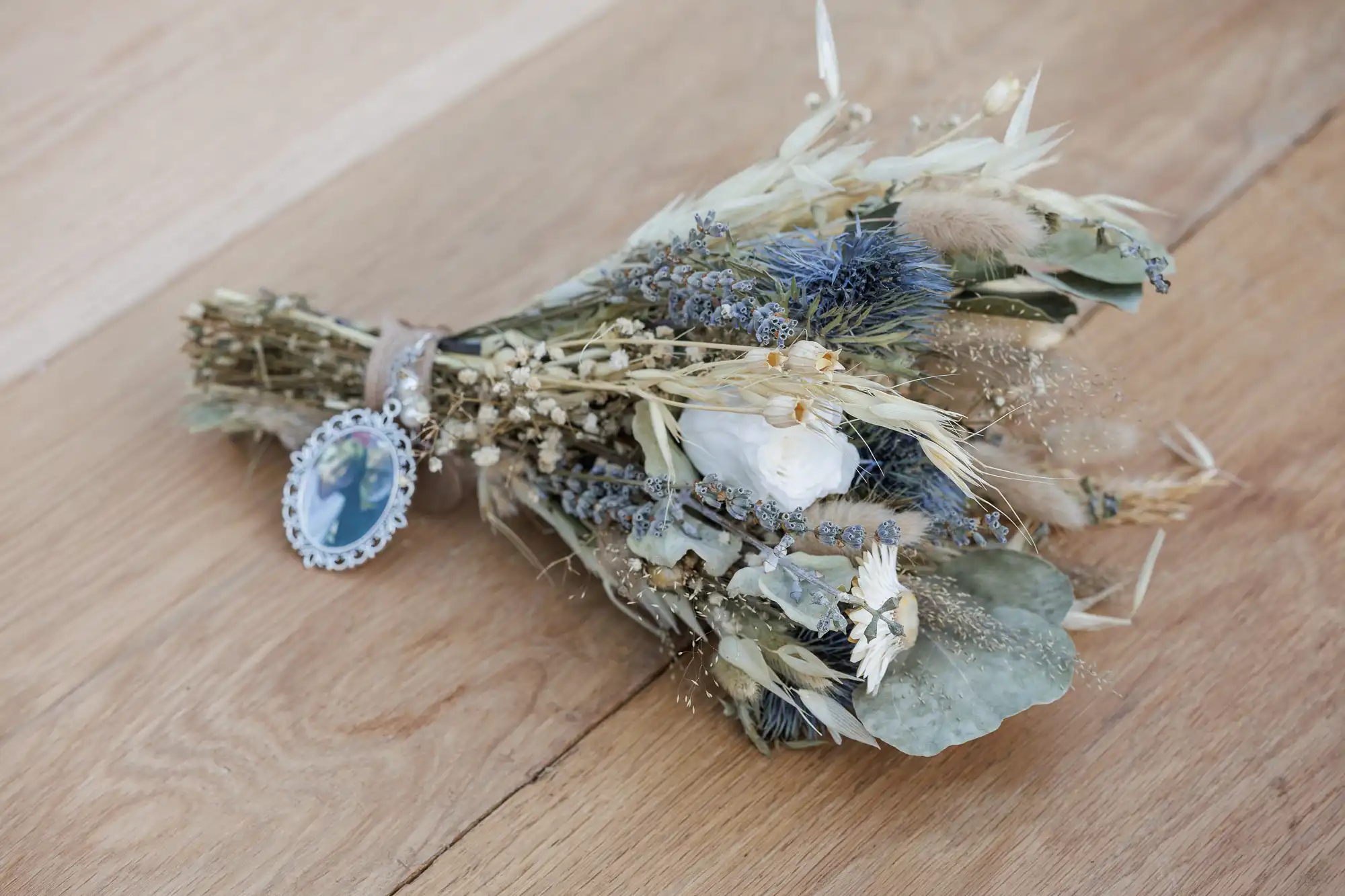 A small posy of dried flowers, including white rose, lavender, and assorted grasses, rests on a wooden surface. The posy is tied with twine and adorned with a round photo charm featuring an image in a vintage-style frame.