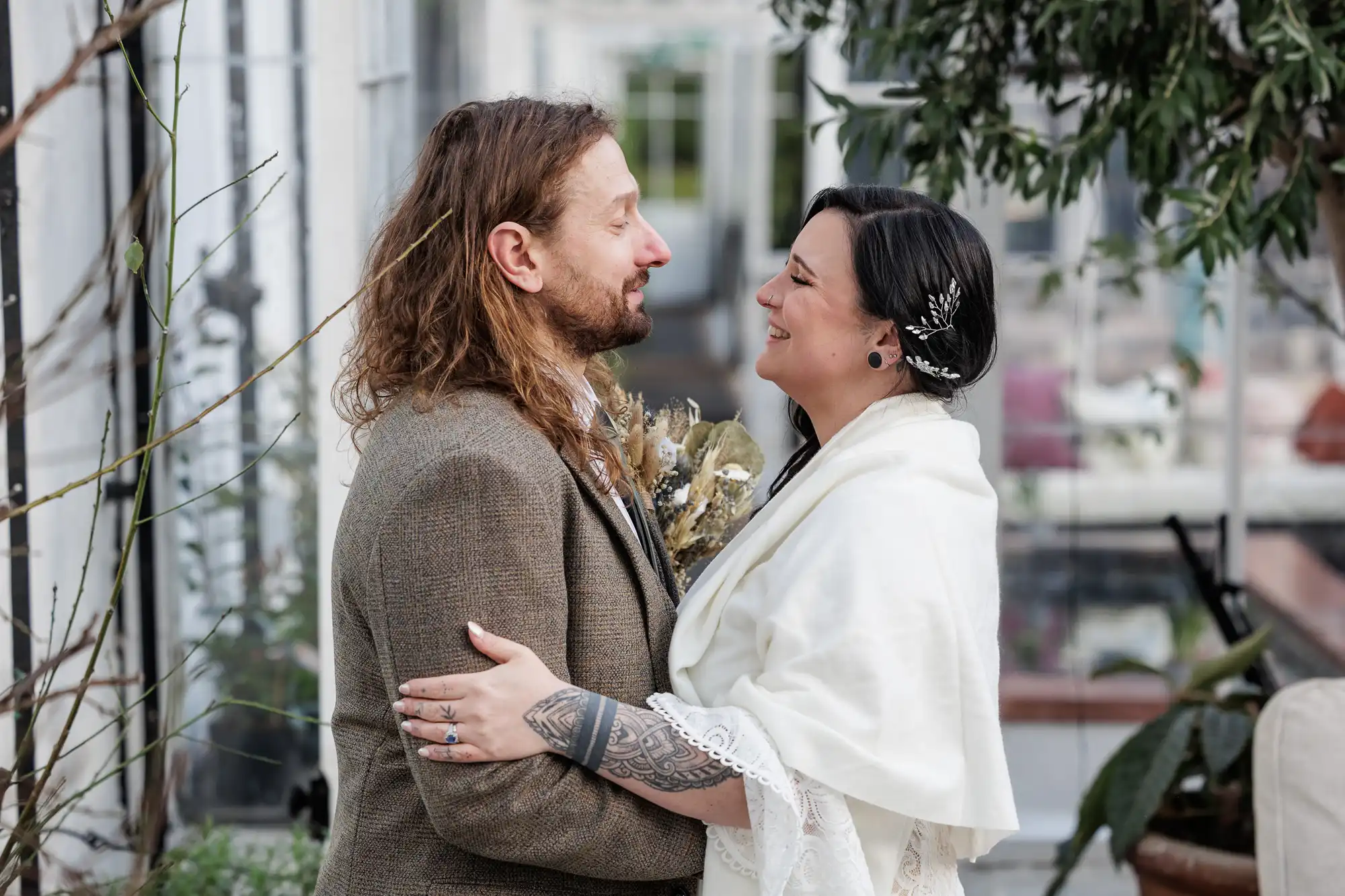 A couple stands indoors, facing each other and smiling warmly. The man has long hair and a beard, wearing a brown suit. The woman wears a white shawl and hairpins, showing a tattooed arm. Green plants and large windows are visible in the background.