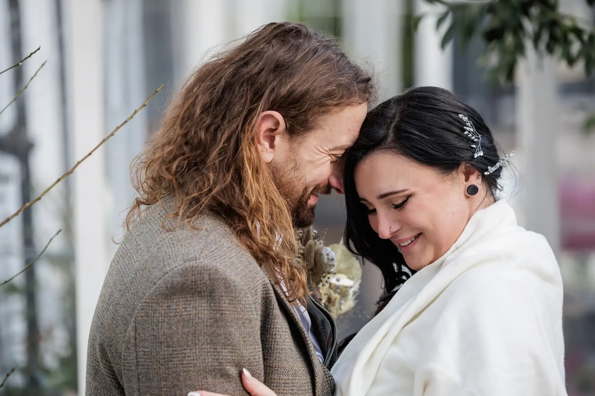 A man with long, wavy brown hair and a beard, dressed in a brown tweed jacket, stands forehead-to-forehead with a smiling woman with long dark hair, draped in a white shawl. They appear joyful and close, sharing an intimate, happy moment outdoors.