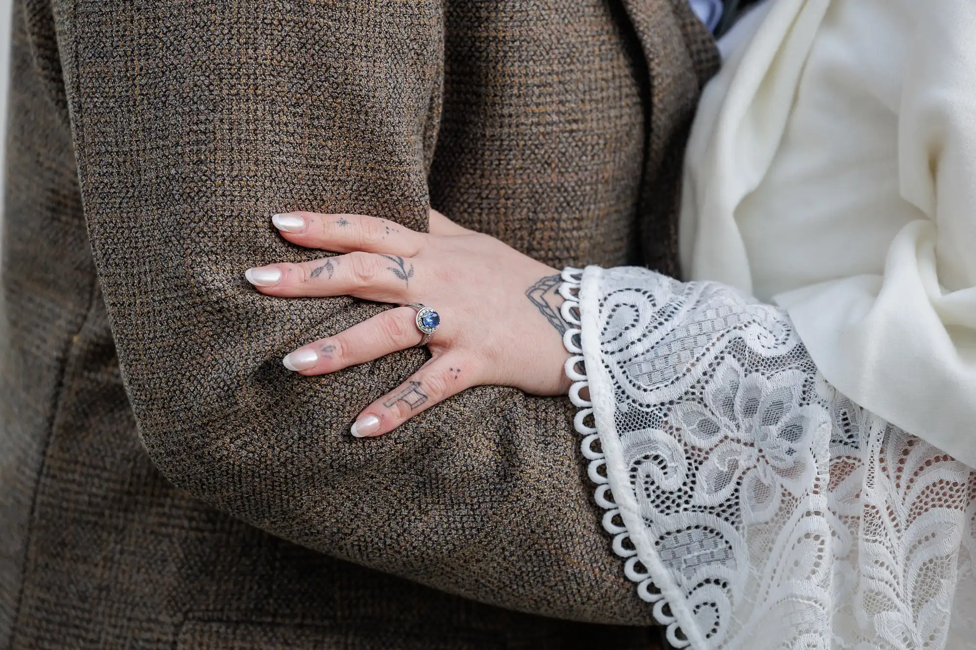 A close-up of two people embracing. One person in a brown tweed jacket is hugged by another wearing a white, lace-detailed sleeve. The latter’s hand, with a blue-stone ring and manicured nails, rests on the other's arm, showing tattoos on their fingers.