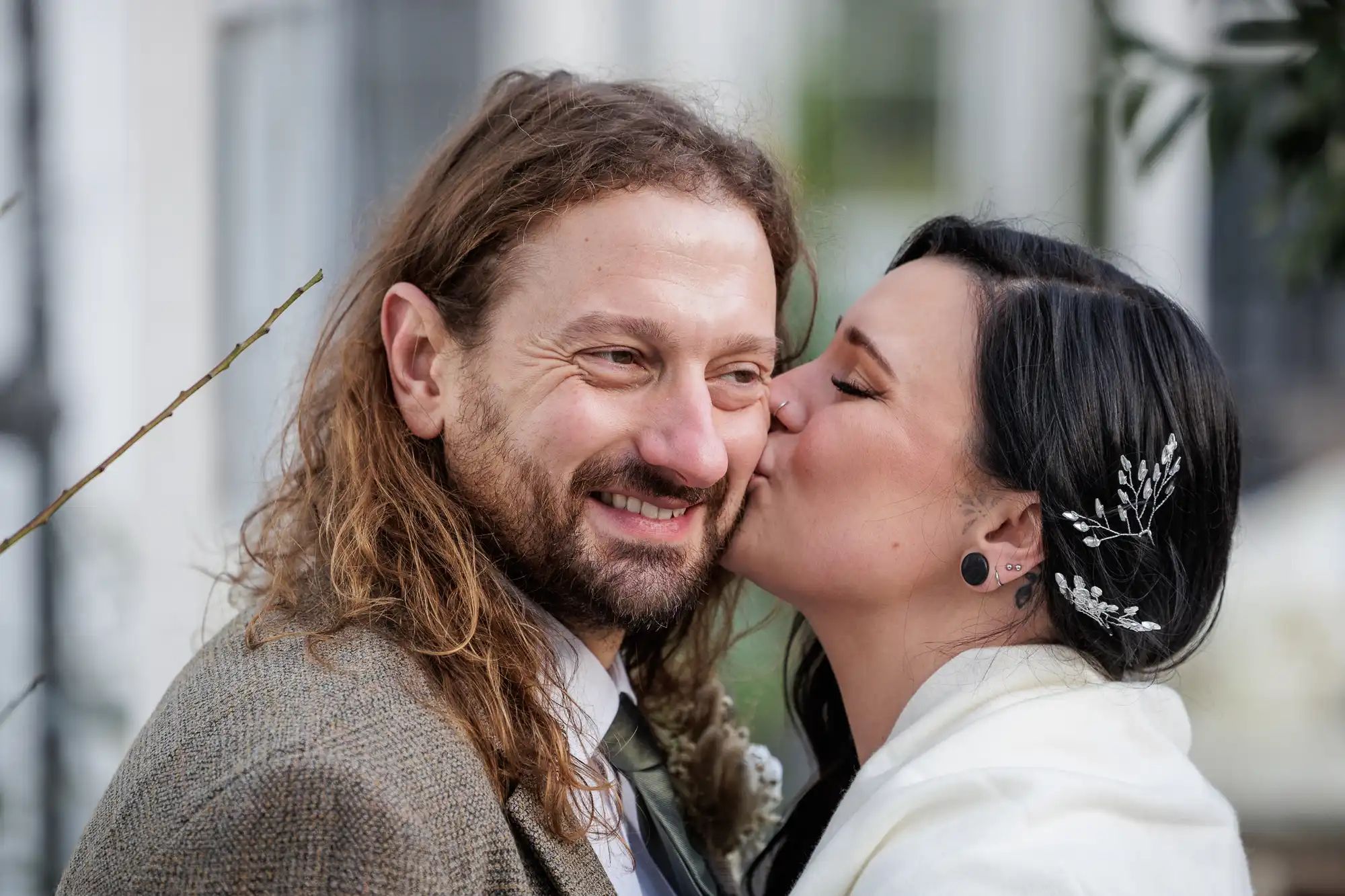 A bride with dark hair, a silver floral hairpin, and a white jacket kisses the cheek of a smiling groom with long brown hair and a beard wearing a brown blazer. Both appear joyful in an outdoor setting.