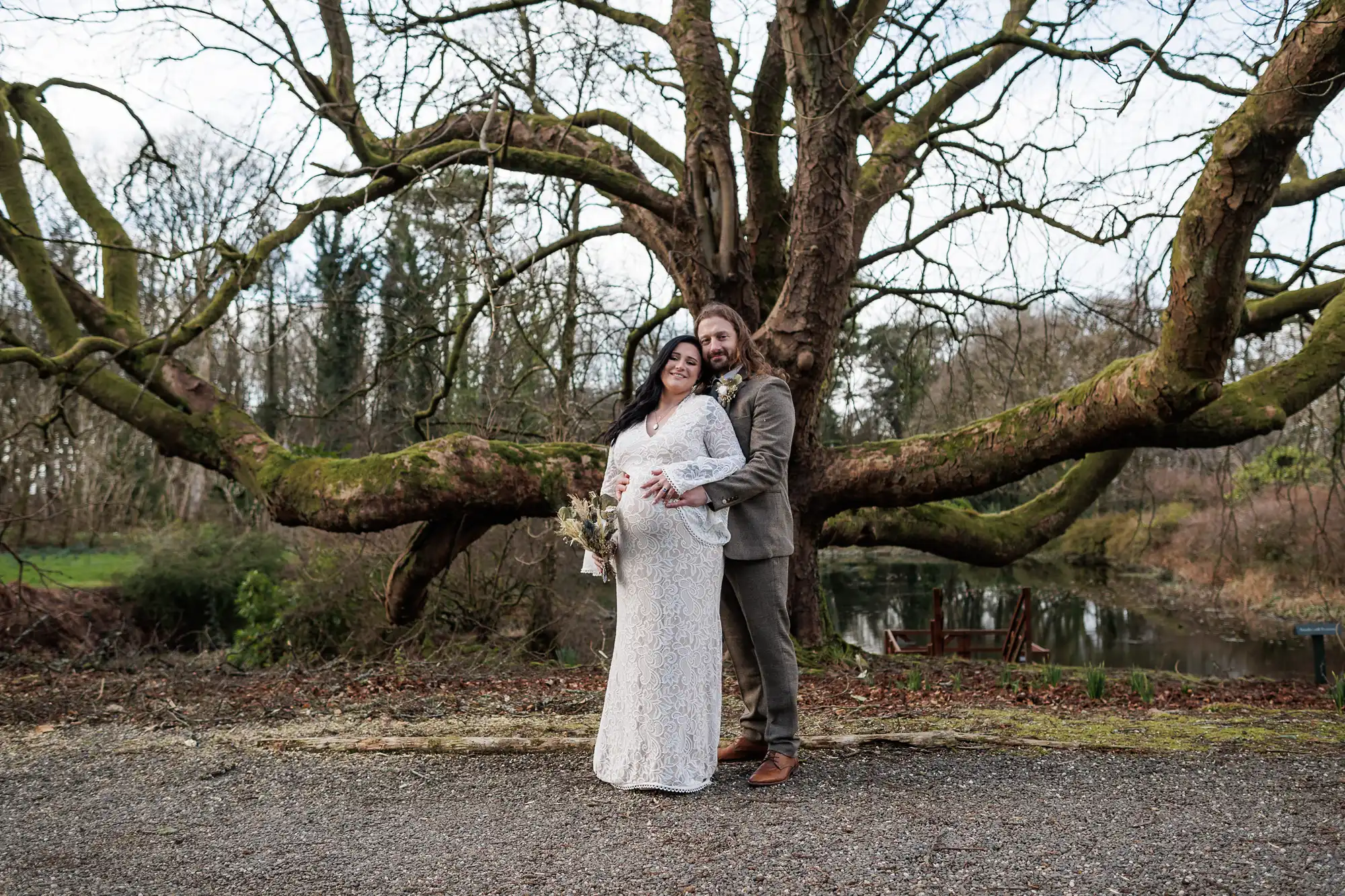 A smiling couple pose outdoors in front of a large, leafless tree with spreading branches. The woman, wearing a white lace dress and holding a bouquet, stands in front of the man, who is dressed in a brown suit and hugging her from behind.