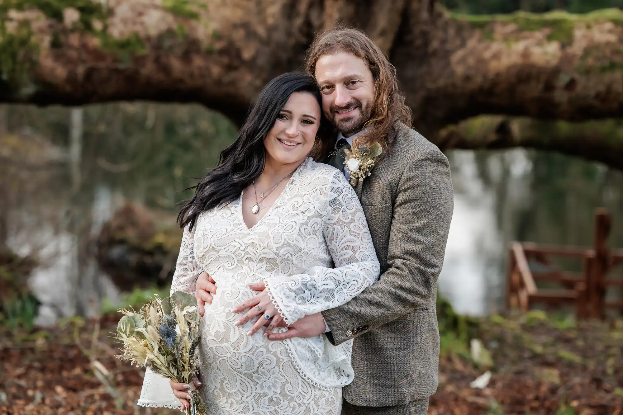 A smiling couple pose outdoors, standing in front of a large tree. The woman, wearing a white patterned dress, is visibly pregnant and holds a dried flower bouquet. The man, in a brown suit, stands close behind her with his hand on her bump.