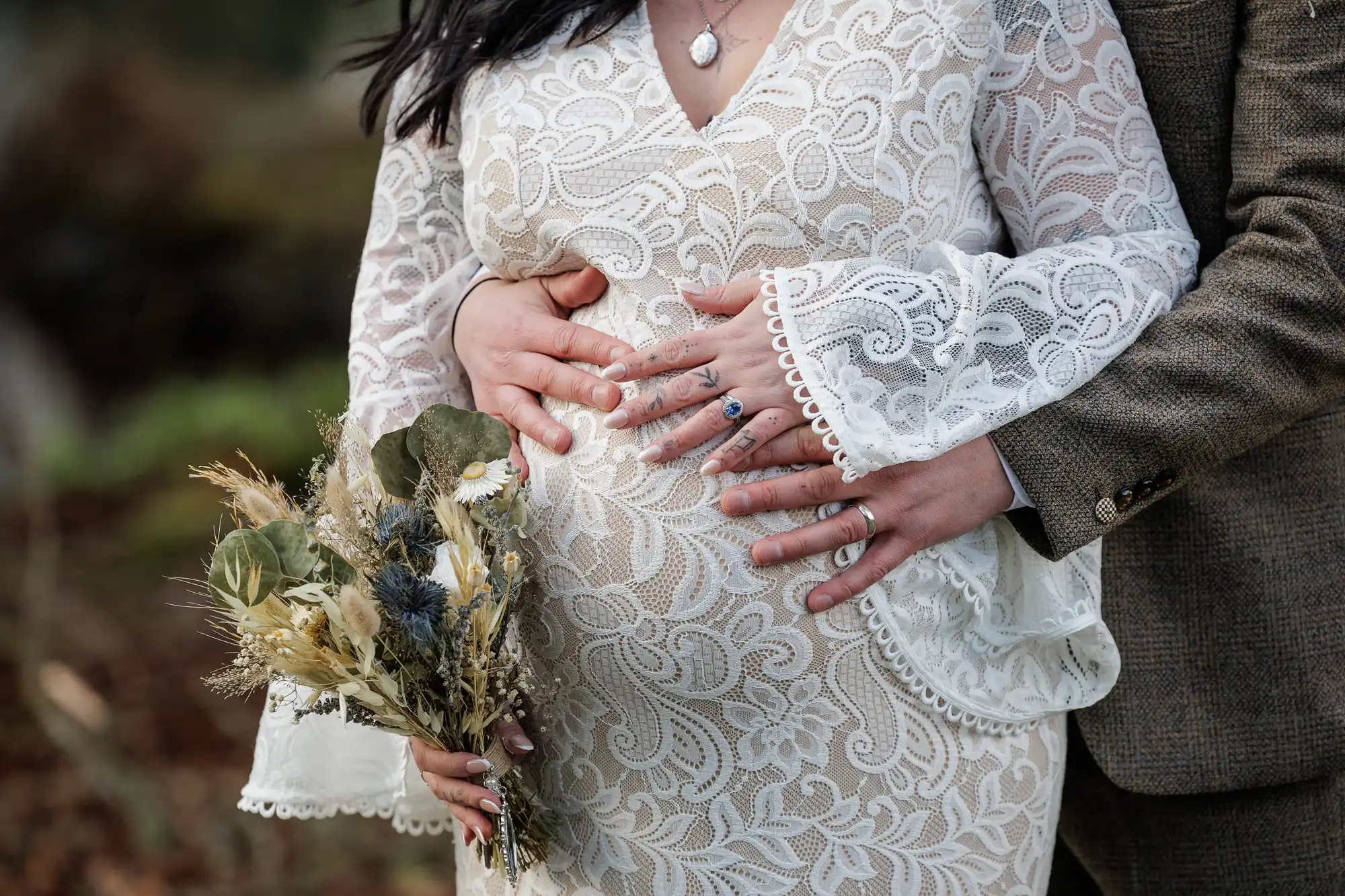 A pregnant woman in a white lace dress holds a bouquet of dried flowers. A person stands behind her, wrapping their hands around her bump. Both wear rings, and only their torsos are visible, suggesting an intimate, outdoor maternity moment.