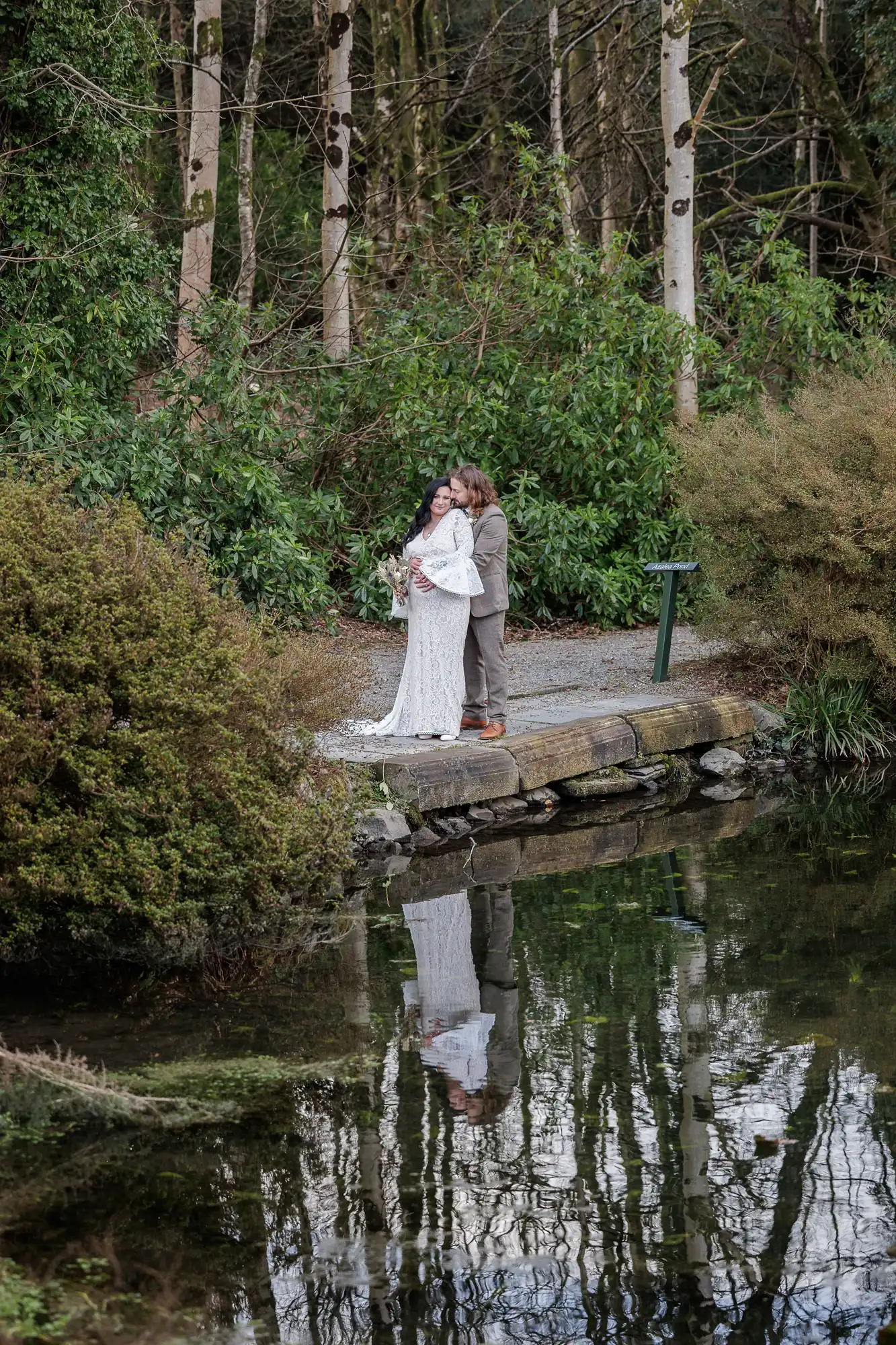 A bride in a white gown and a groom in a tan suit stand close together on a stone path by a pond, surrounded by greenery and trees. Their reflection appears in the water, and the background features dense foliage and tall, thin tree trunks.
