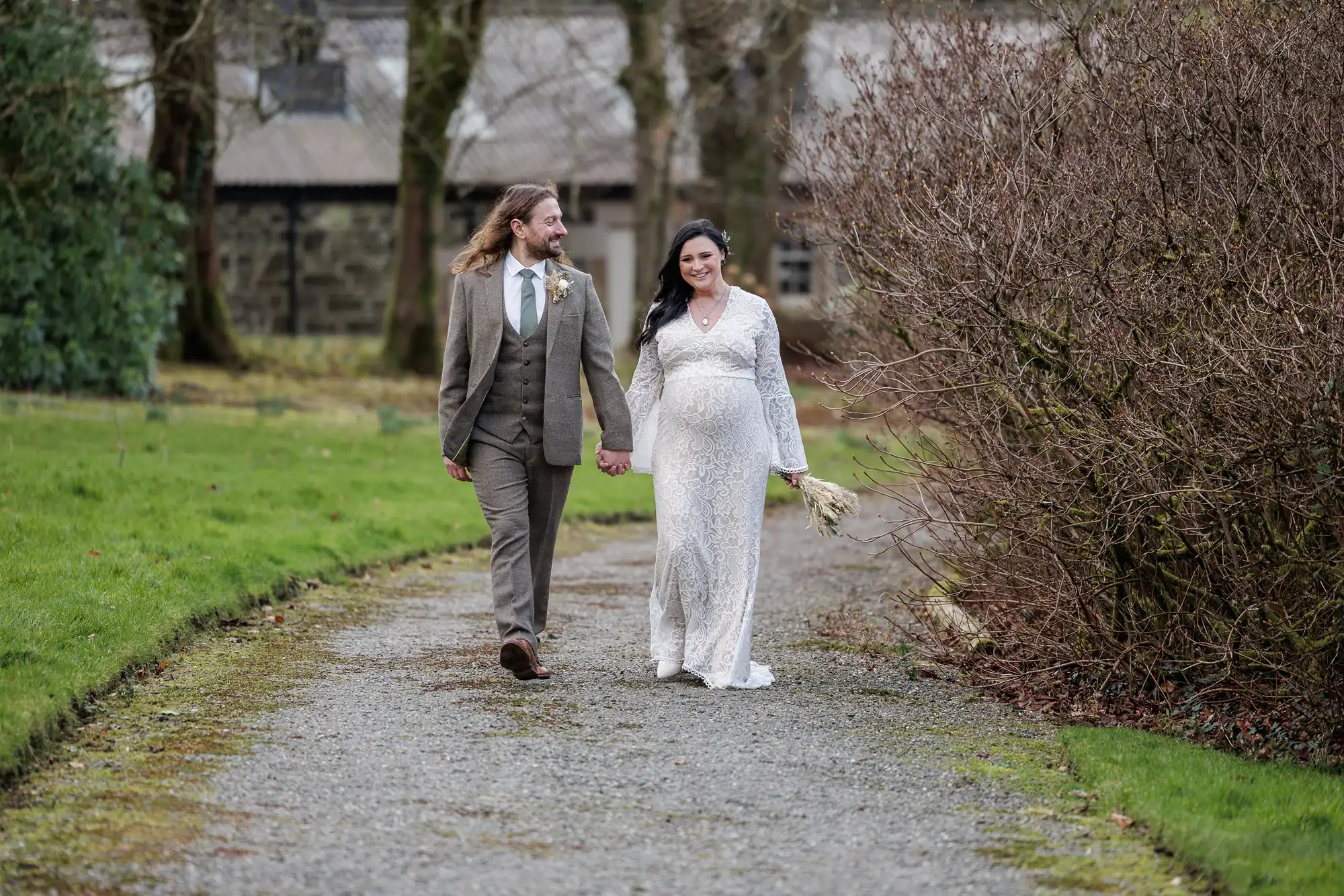 A smiling couple walk hand in hand down a gravel path. The man wears a brown suit and tie, and the woman, who appears pregnant, wears a long white lace dress and holds a small bouquet. Bare bushes and green grass line the path, with buildings in the background.