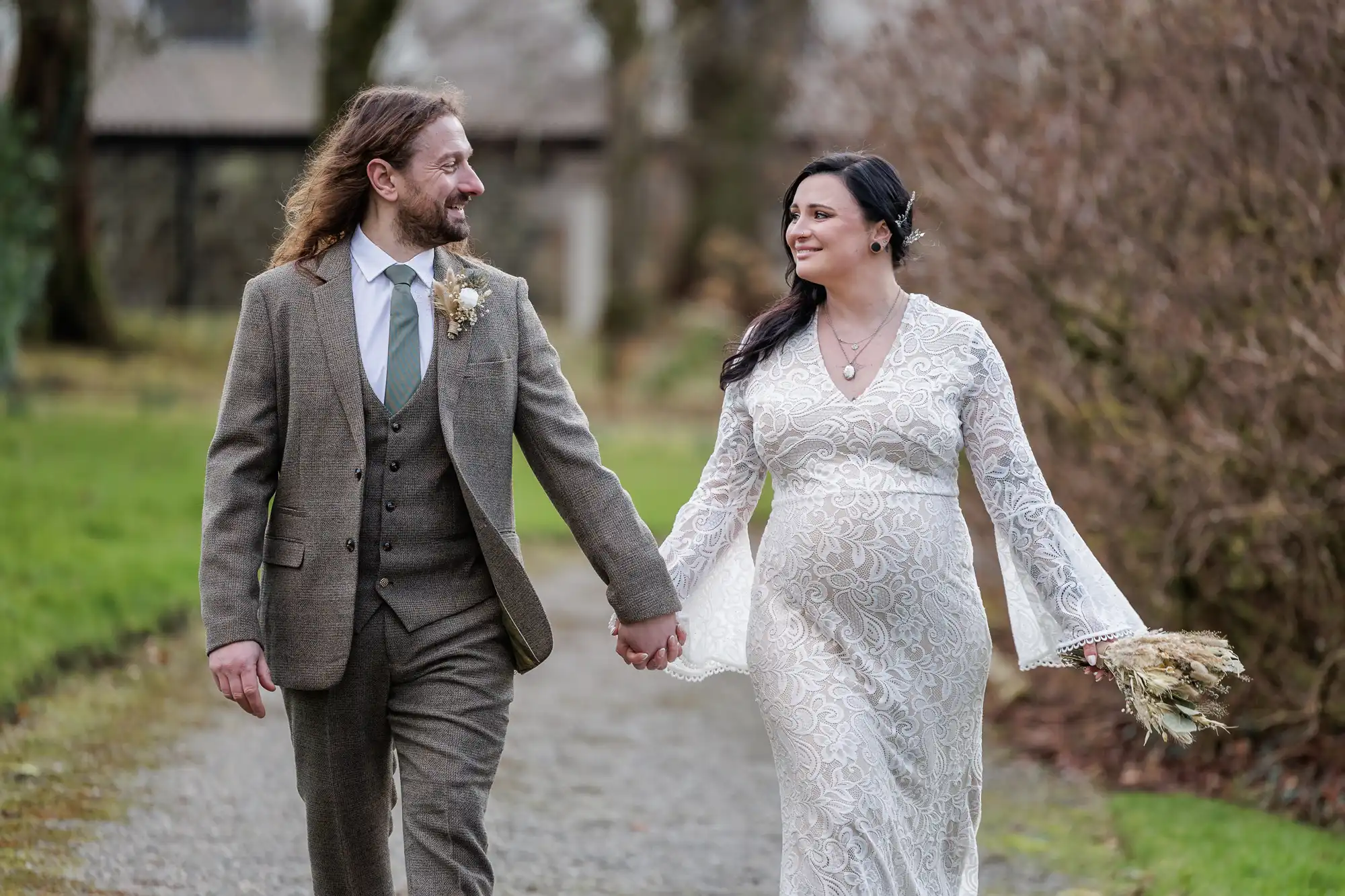 A couple walk hand in hand outdoors on a gravel path. The man wears a brown suit with a waistcoat and tie, and the woman wears a white lace dress, holding a bouquet of dried flowers. Both are smiling and looking at each other, surrounded by greenery and leafless bushes.