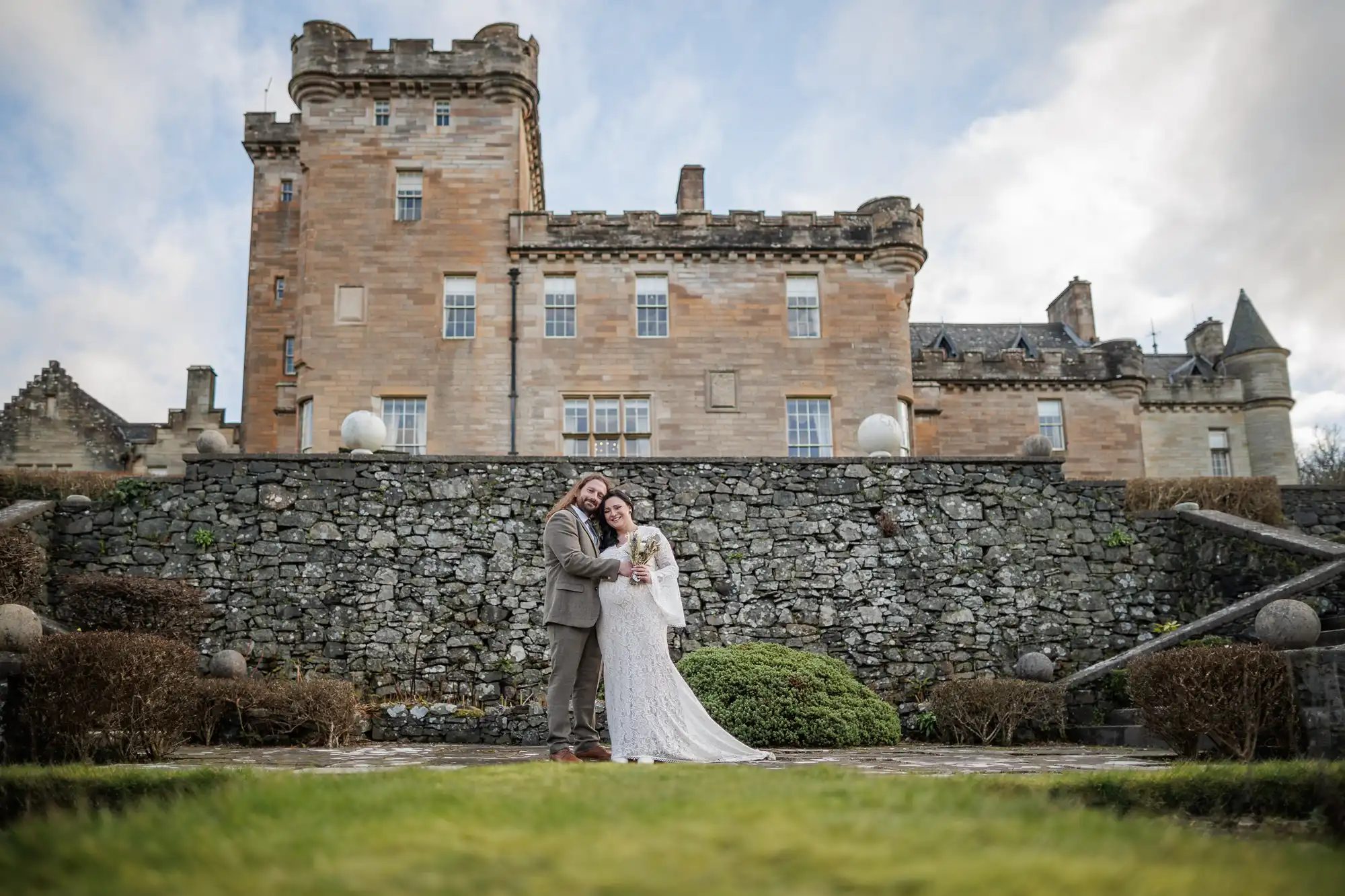 A couple in wedding attire stand closely together, smiling, in front of a large historic stone castle with turrets. The bride holds a bouquet; trimmed bushes and stone steps lead up to the castle under a cloudy sky.