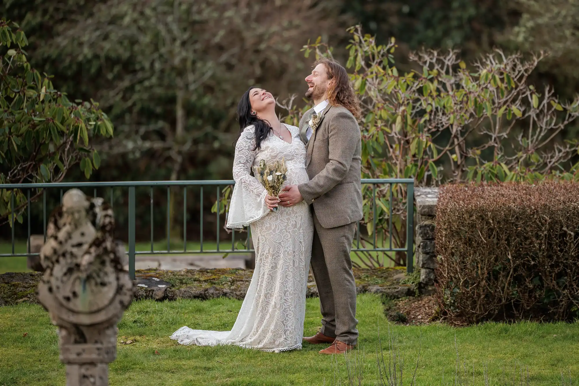 A bride in a white lace gown and a groom in a brown suit stand laughing together outdoors. The bride holds a bouquet as they embrace, both looking joyful. Behind them is a metal railing, shrubs, and greenery in a garden setting.