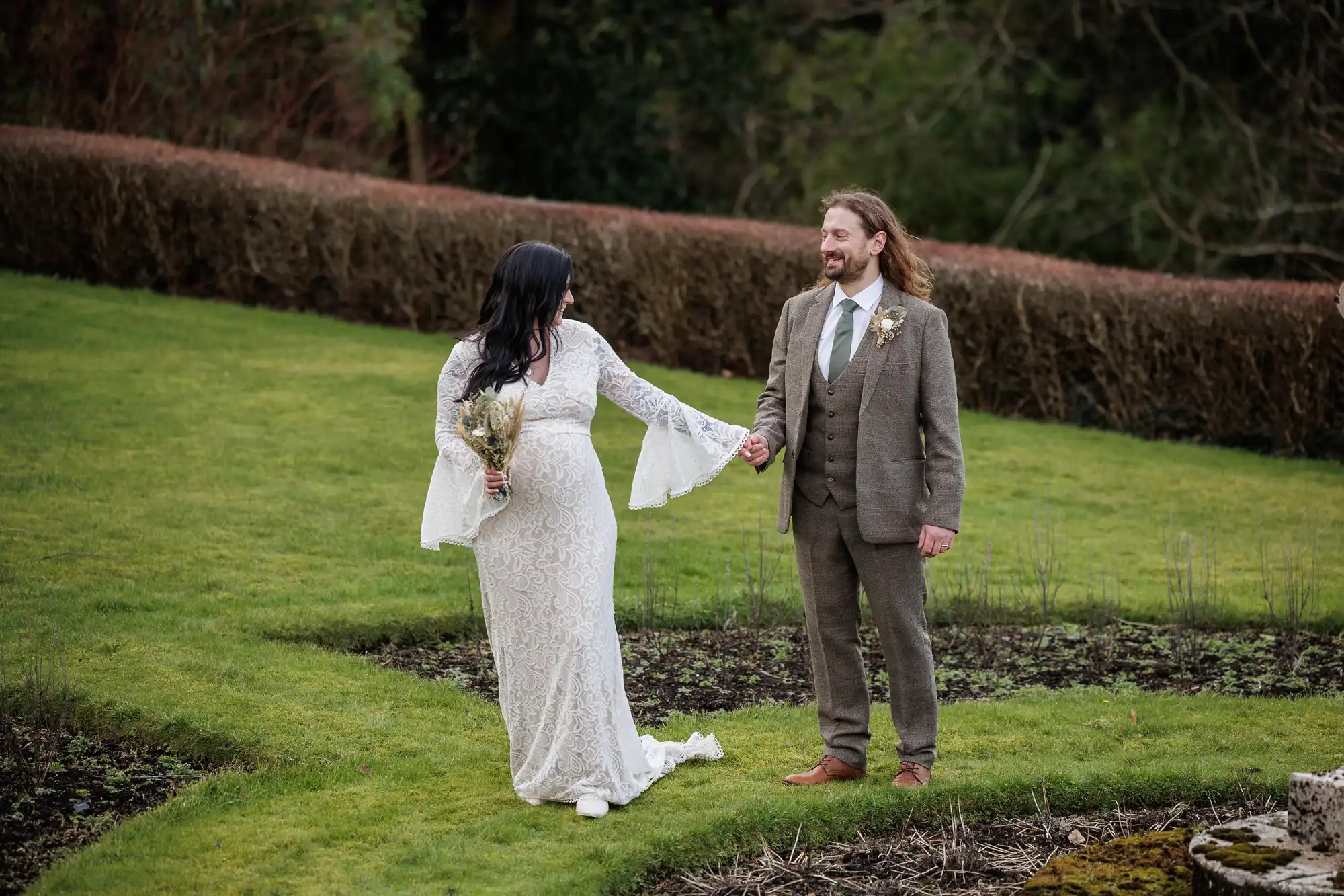A bride in a long white lace dress holding a bouquet and a groom in a brown suit with a buttonhole smile at each other whilst holding hands, standing on green grass in a garden with trimmed hedges and trees in the background.
