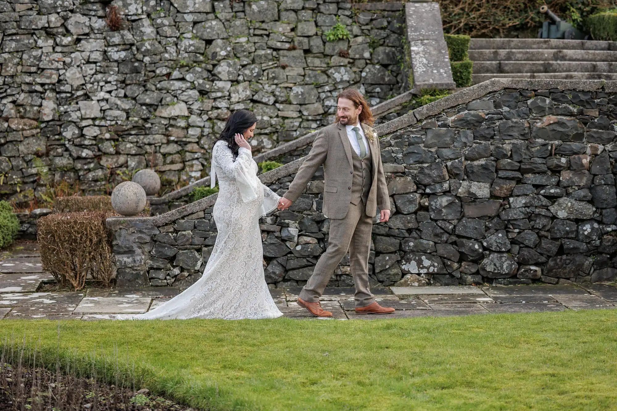 A bride in a long, white lace dress holds hands with a groom in a tan suit as they walk outside along a stone path, with a rustic stone wall and steps in the background, and green grass in the foreground. Both appear happy and smiling.