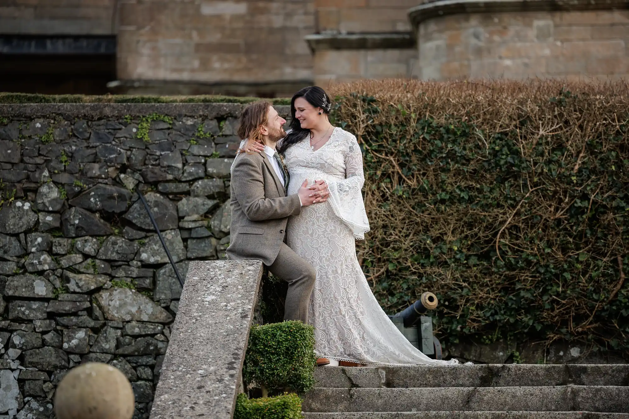 A couple pose on outdoor stone steps. The groom in a brown suit sits, gazing lovingly at the bride, who stands in a long, white lace dress, holding her pregnant bump. A stone wall and trimmed hedge are in the background, suggesting a rustic setting.