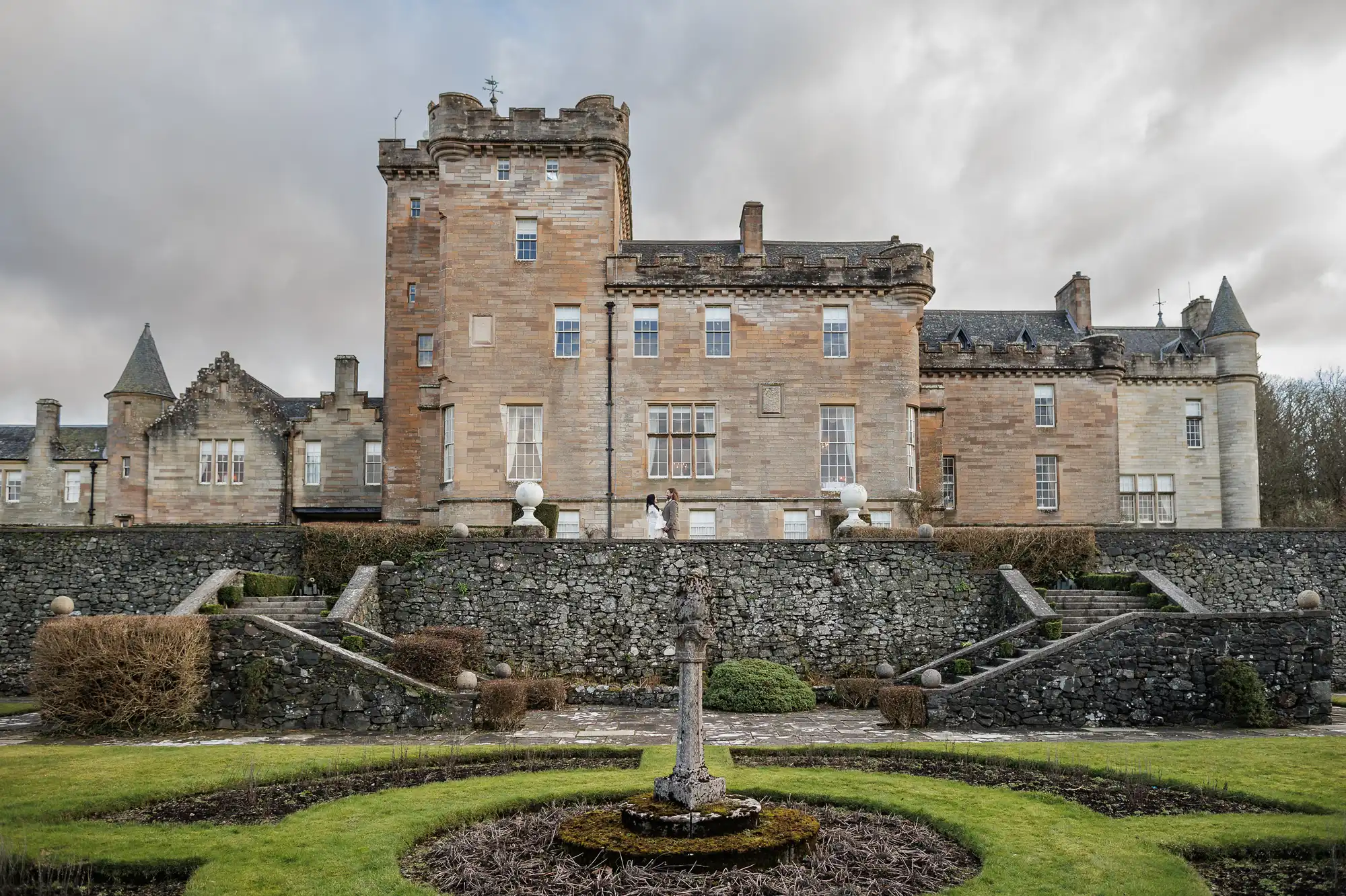 A large stone castle with turrets and battlements sits under a cloudy sky. In front, symmetrical terraced gardens feature trimmed bushes, stone steps, and a weathered stone fountain at the centre of a grassy lawn.