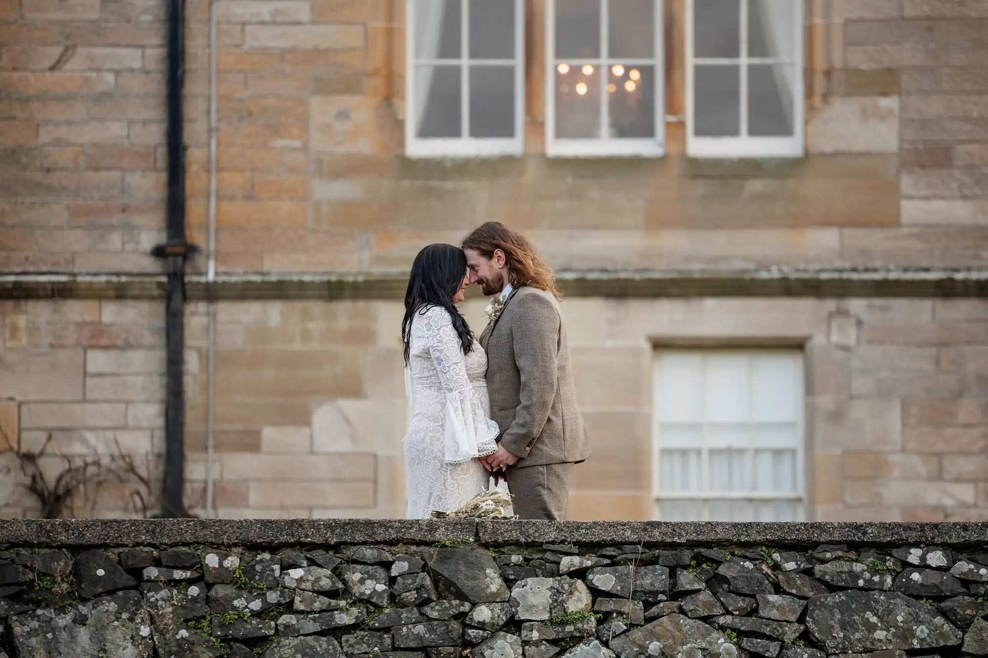 A couple stands face to face, gently touching foreheads and holding hands. The woman wears a white lace dress, and the man wears a tan suit. They stand on a stone terrace with a large brick building and tall windows in the background.