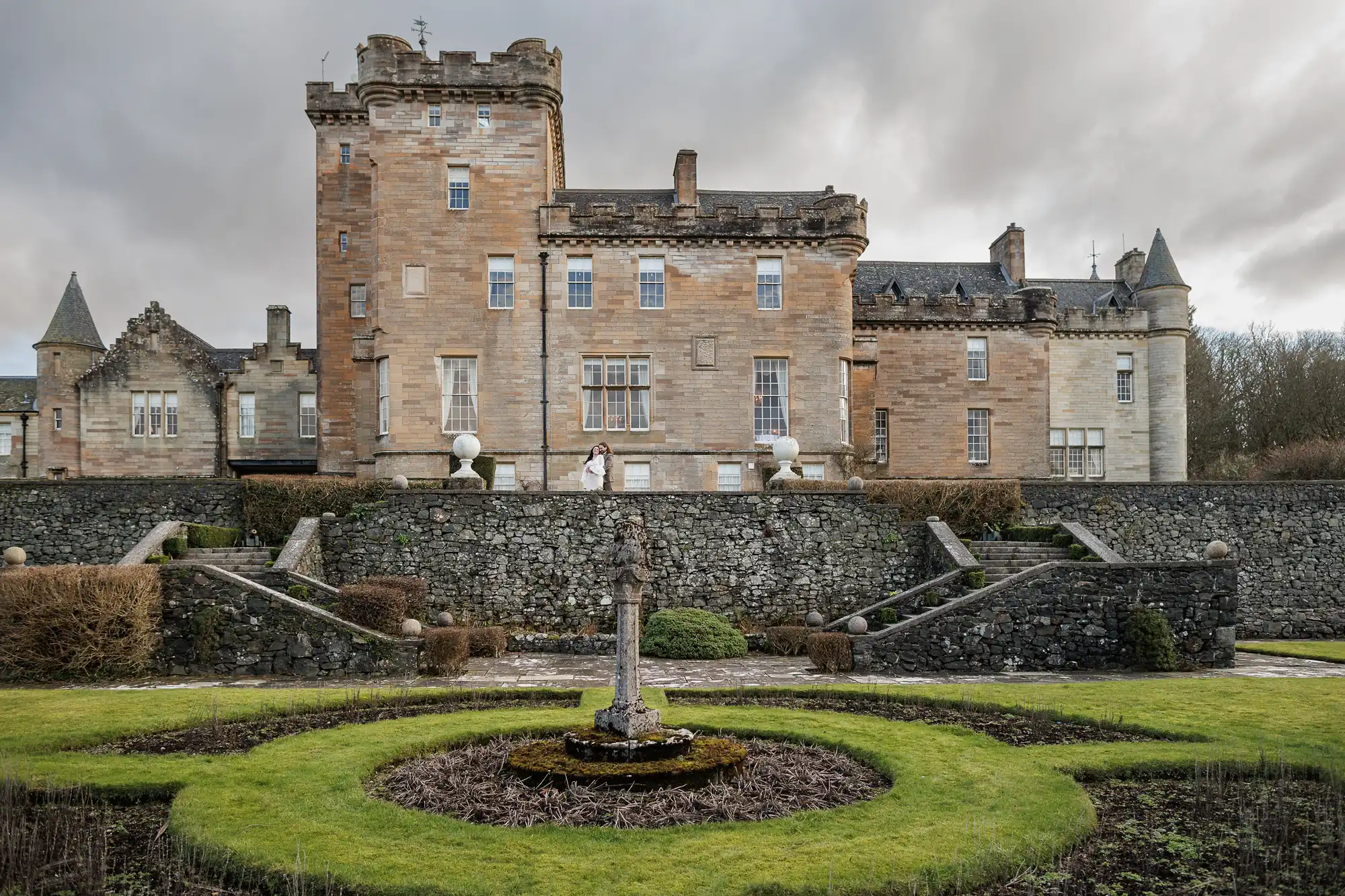 A large historic stone castle with towers and crenellations stands behind terraced stone walls and a formal garden featuring a circular fountain at the centre. Bushes and trimmed lawn frame the fountain under a cloudy sky.
