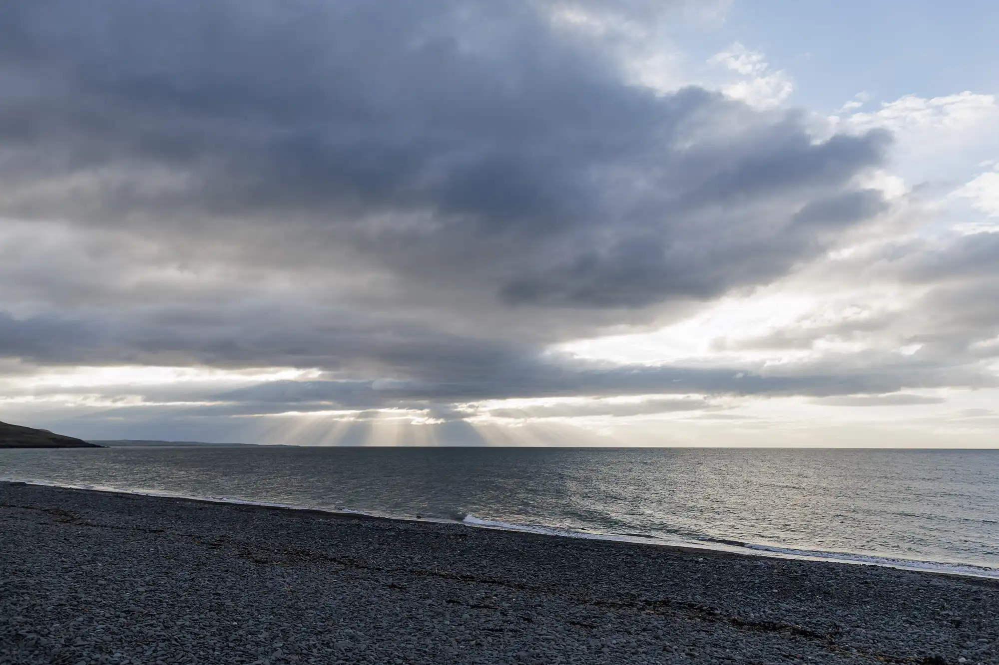 A rocky beach stretches along the bottom of the image, meeting a calm sea. Above, dramatic clouds partially cover the sky, with rays of sunlight breaking through and shining down onto the water, creating a peaceful and moody scene.
