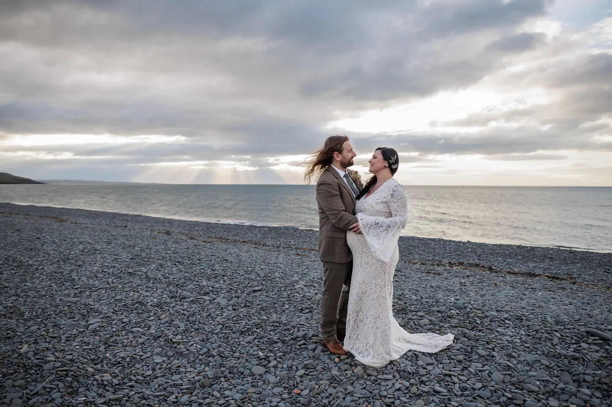 A couple in wedding attire stands on a rocky beach at sunset. The groom wears a brown suit, and the bride wears a white lace dress. They hold each other and smile, with the dramatic sky, sea, and beams of sunlight in the background.