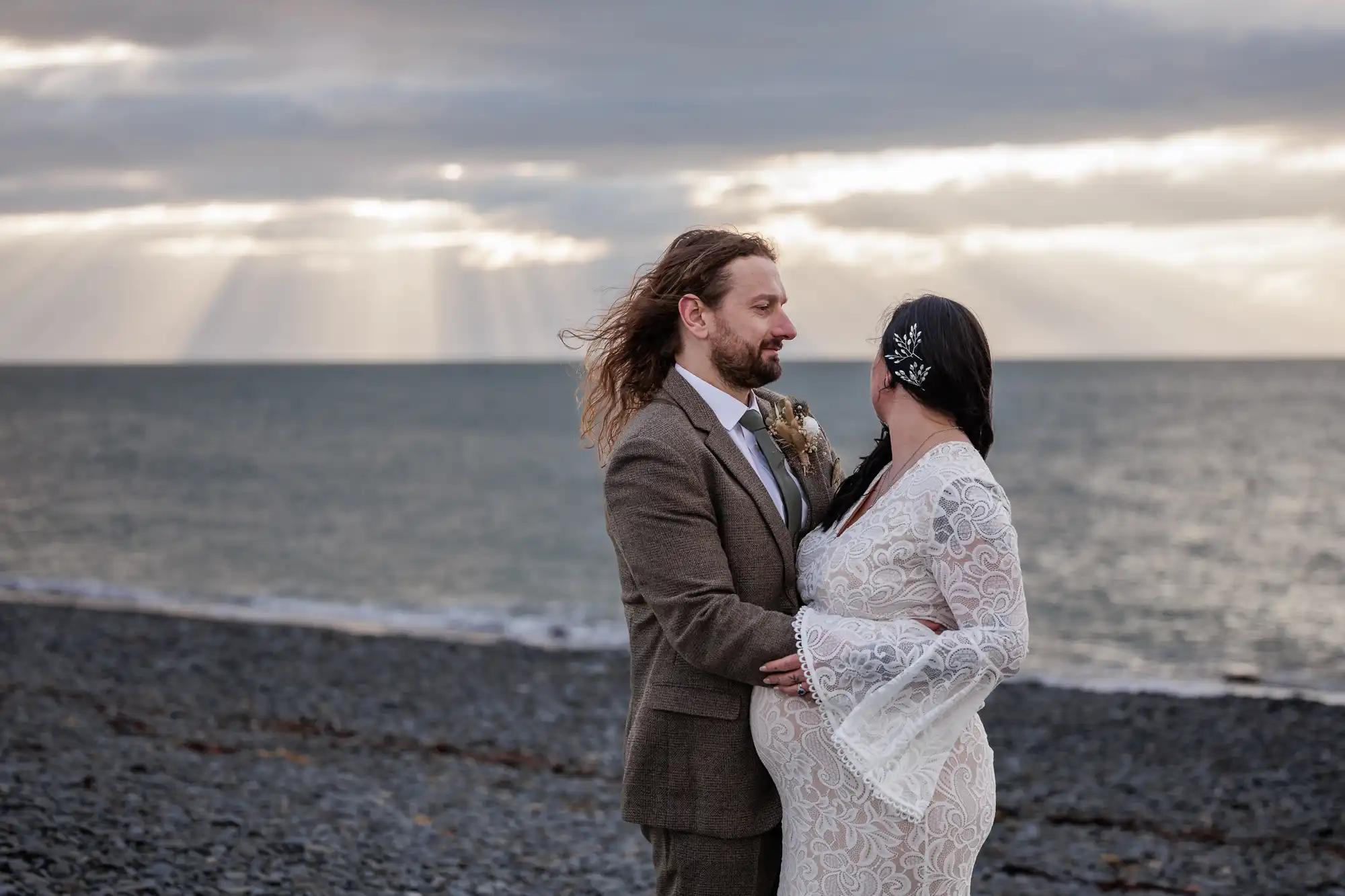 A couple stands on a rocky beach at sunset. The man, in a brown suit, faces a woman in a long white lace dress with flared sleeves. They embrace tenderly, with the calm sea and dramatic sun rays through clouds in the background.