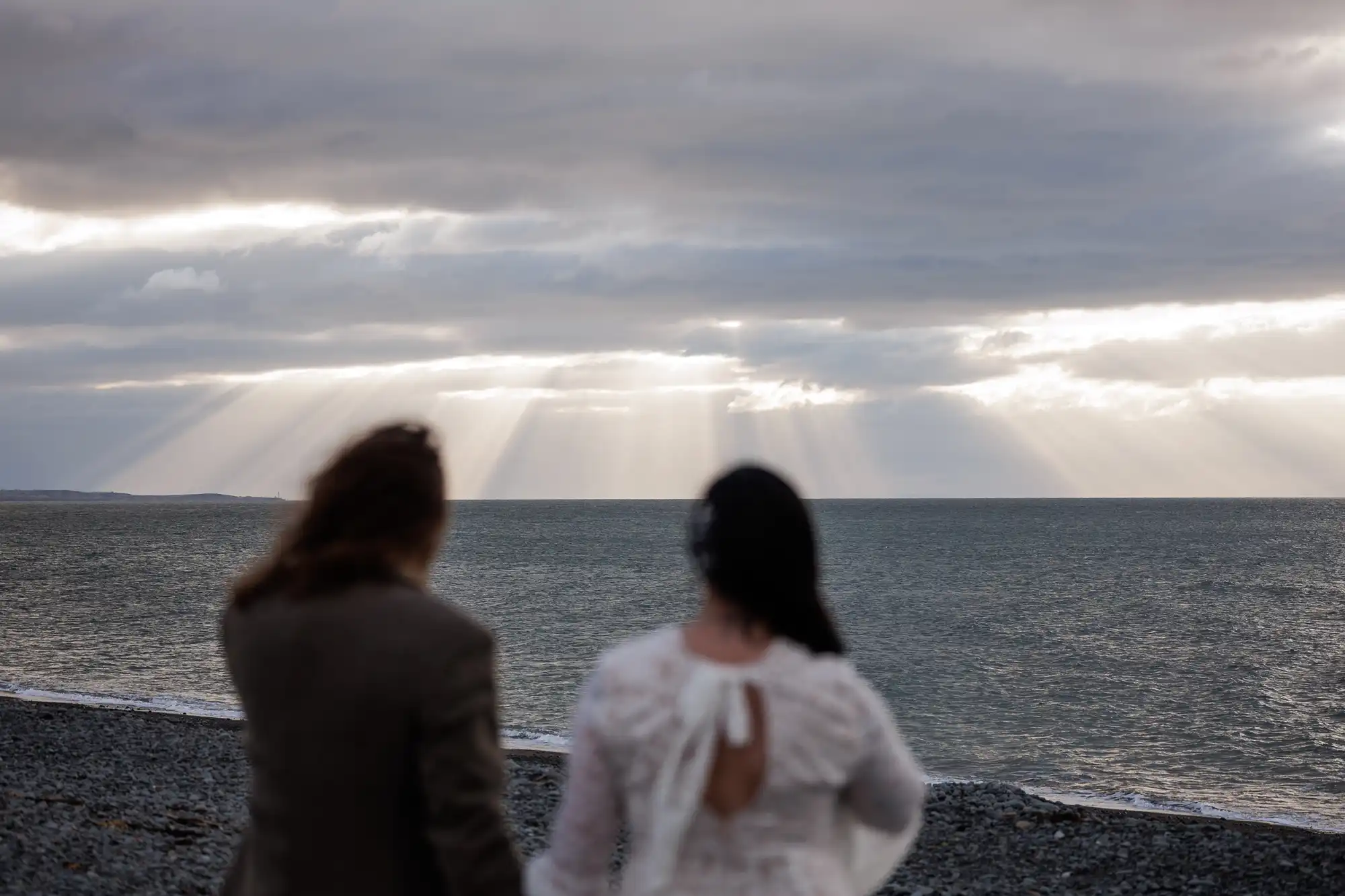 Two people, one in a white dress and one in a brown jacket, stand on a rocky beach facing the sea. Sunbeams break through dramatic clouds over the water, casting light onto the sea and creating a serene, contemplative atmosphere.