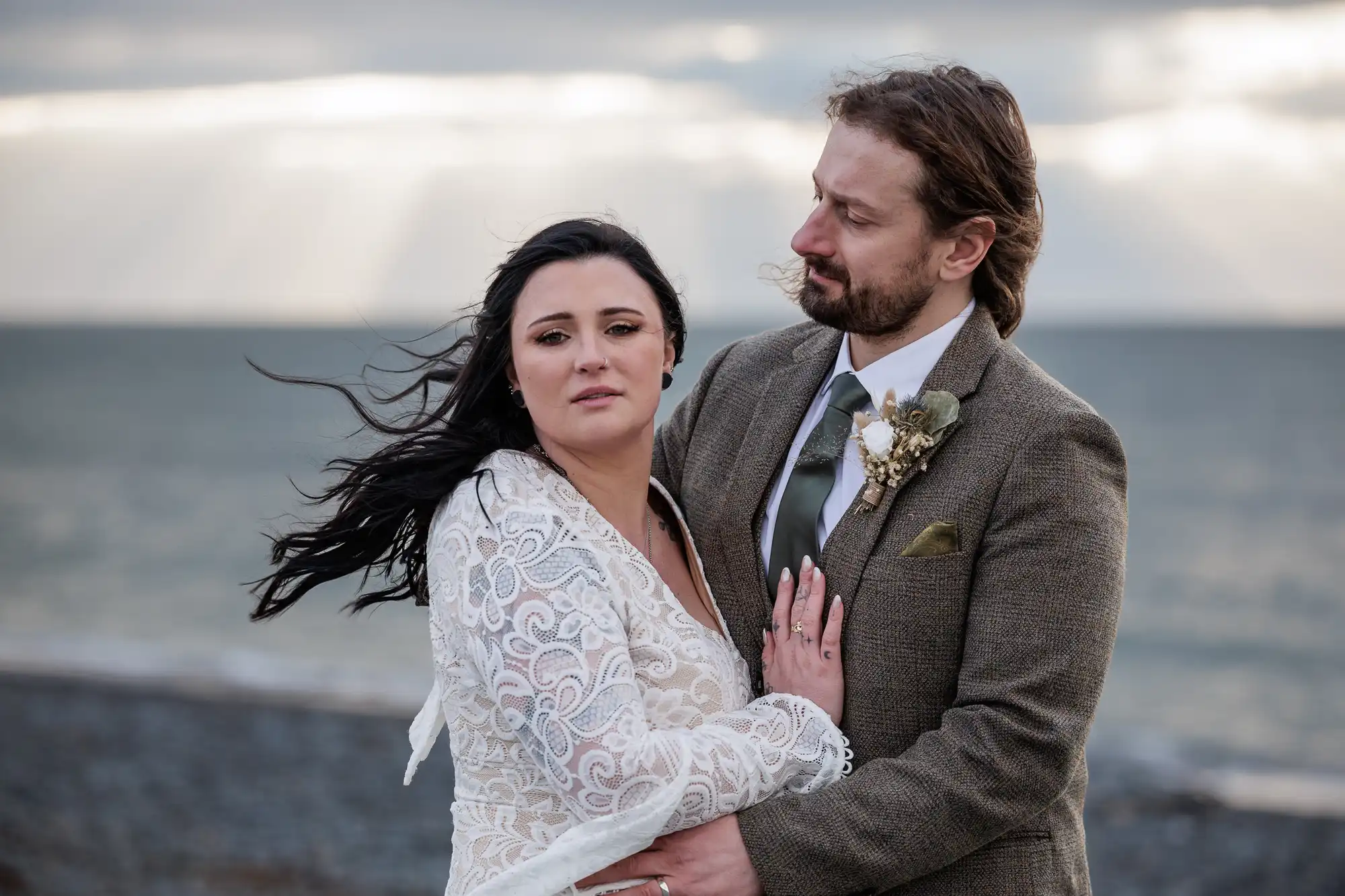 A bride in a white lace dress and a groom in a brown suit stand close together on a rocky beach. The groom holds the bride as she looks at the camera, her dark hair windswept. He gazes at her lovingly. The sea and a cloudy sky are in the background.
