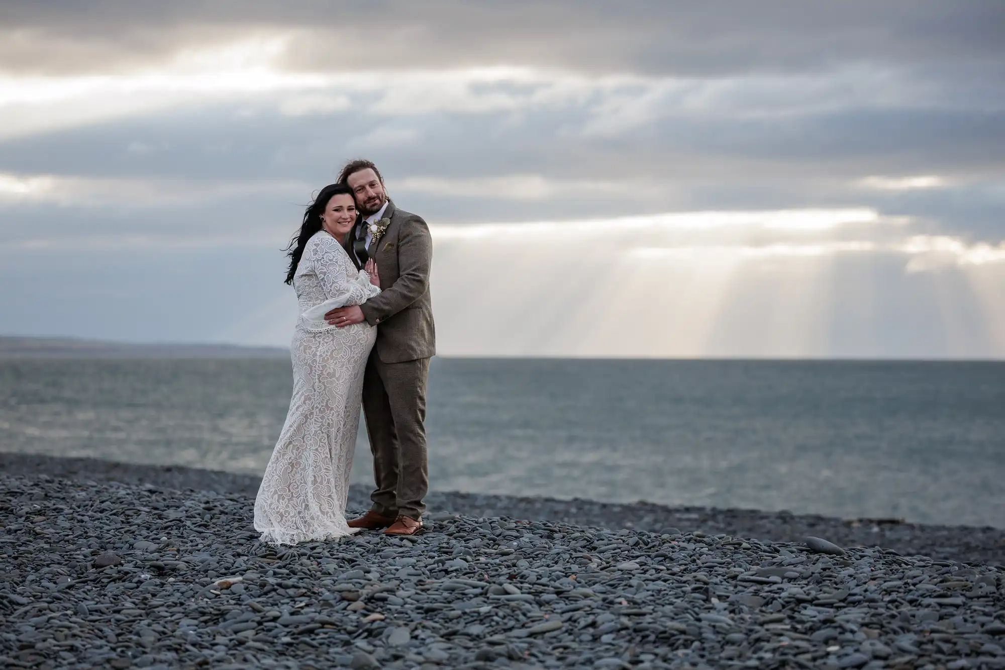 Glenapp Castle wedding photographer photo of a bride in a white lace dress and a groom in a taupe suit stand arm-in-arm on a rocky beach. They smile at the camera, with the sea and dramatic, cloudy sky behind them. Sun rays stream through the clouds, creating a serene, romantic scene.