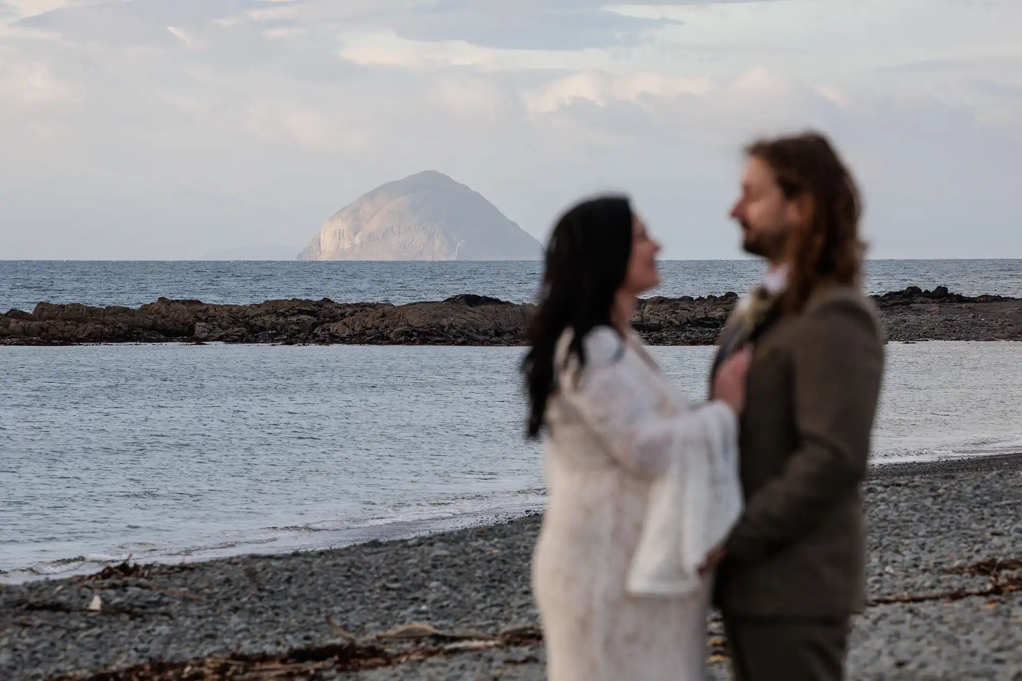 A blurred couple stands close together on a rocky beach in the foreground, facing each other. In the distance, an island rises from the sea under a cloudy sky. The focus is on the scenic background, not the couple.
