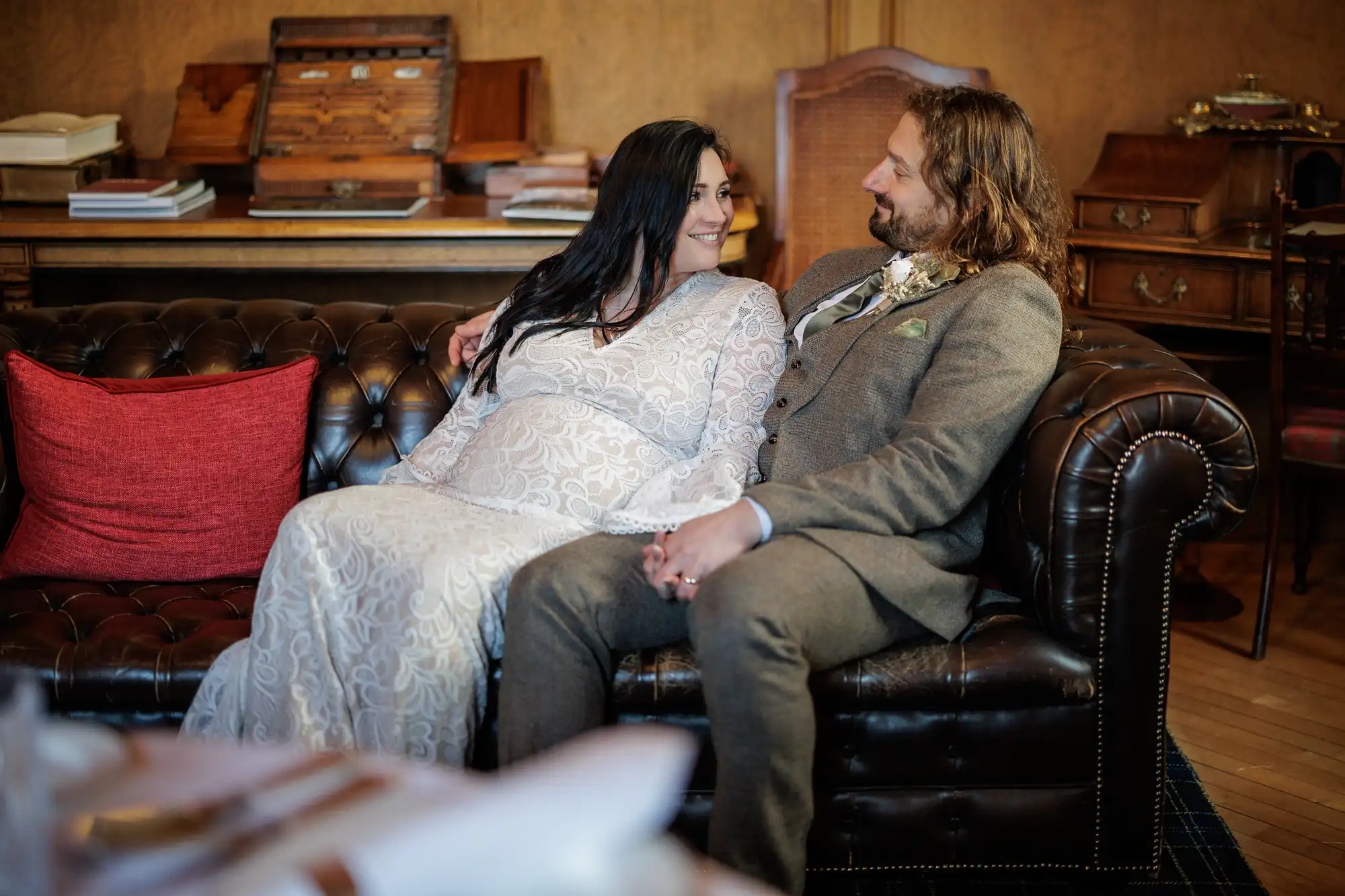 A couple in wedding attire sits on a dark leather sofa. The woman wears a white lace dress and the man wears a brown suit. They hold hands and smile at each other. The room has wooden furniture, books, and a red cushion on the sofa.
