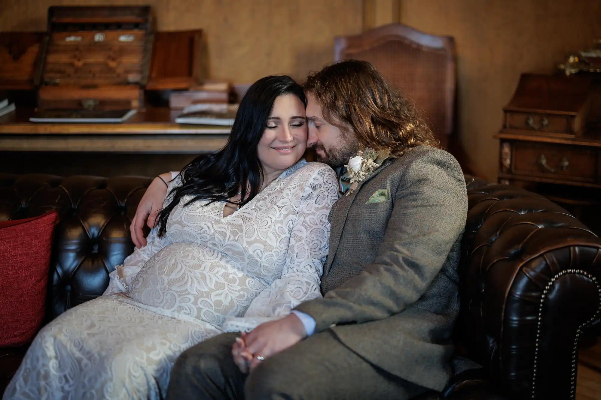 A couple in wedding attire sit close together on a brown leather sofa, eyes closed and smiling, with the woman wearing a white lace dress and the man in a brown suit with a buttonhole. They gently touch foreheads, holding hands in a warm, intimate moment.