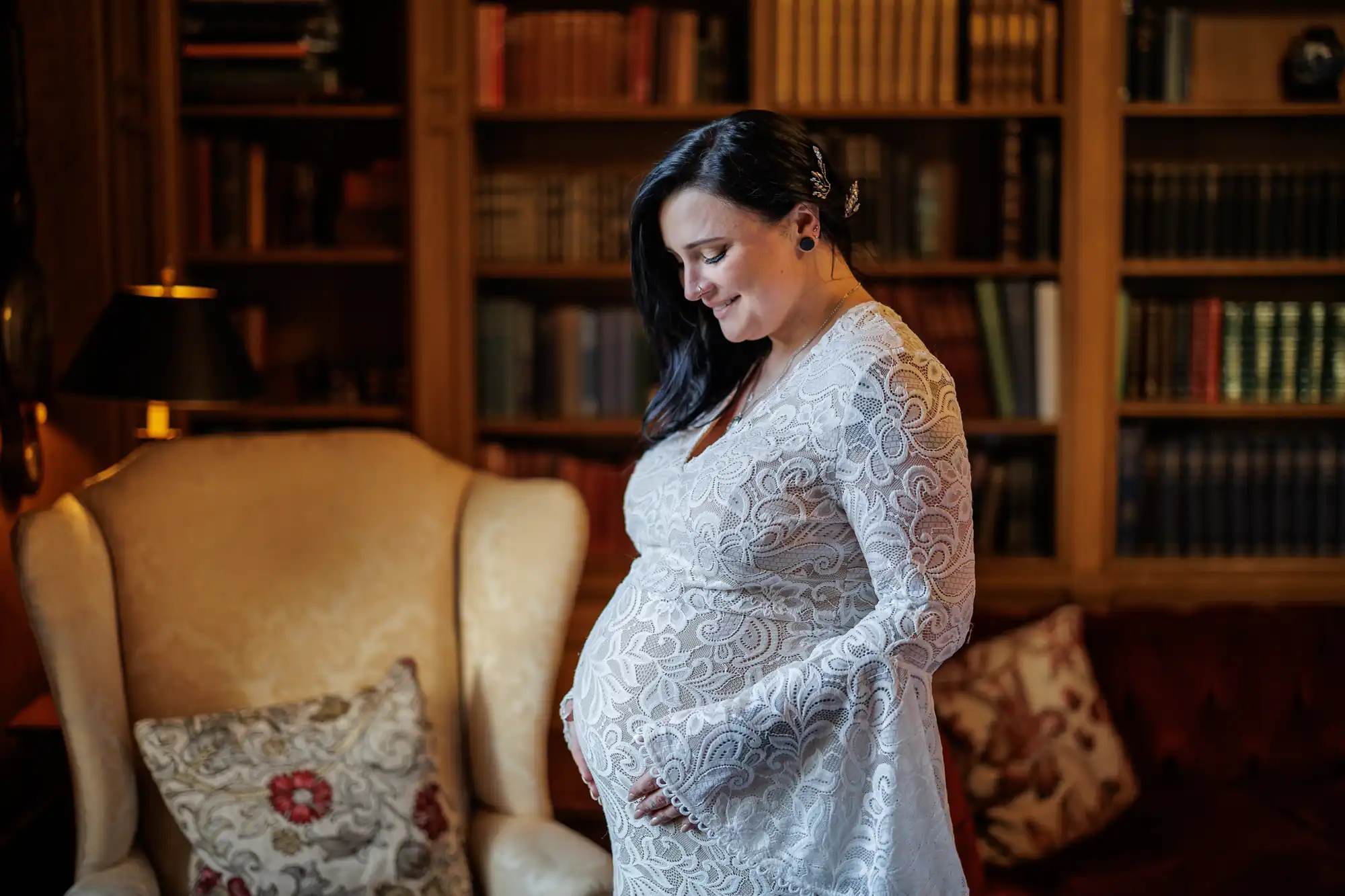 A pregnant woman in a lace dress stands in a cosy library, smiling and looking down at her belly. Bookshelves filled with books line the background, and an armchair with a floral cushion sits nearby. Warm light creates a calm, intimate atmosphere.