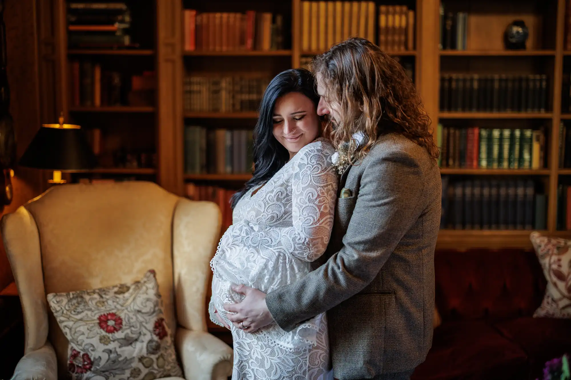 A man embraces a pregnant woman from behind in a cosy, elegant library. The woman wears a white lace dress and smiles softly, resting her hands on her tummy. The man, in a grey suit, gently holds her bump. Bookcases and plush furniture fill the warm, inviting room.