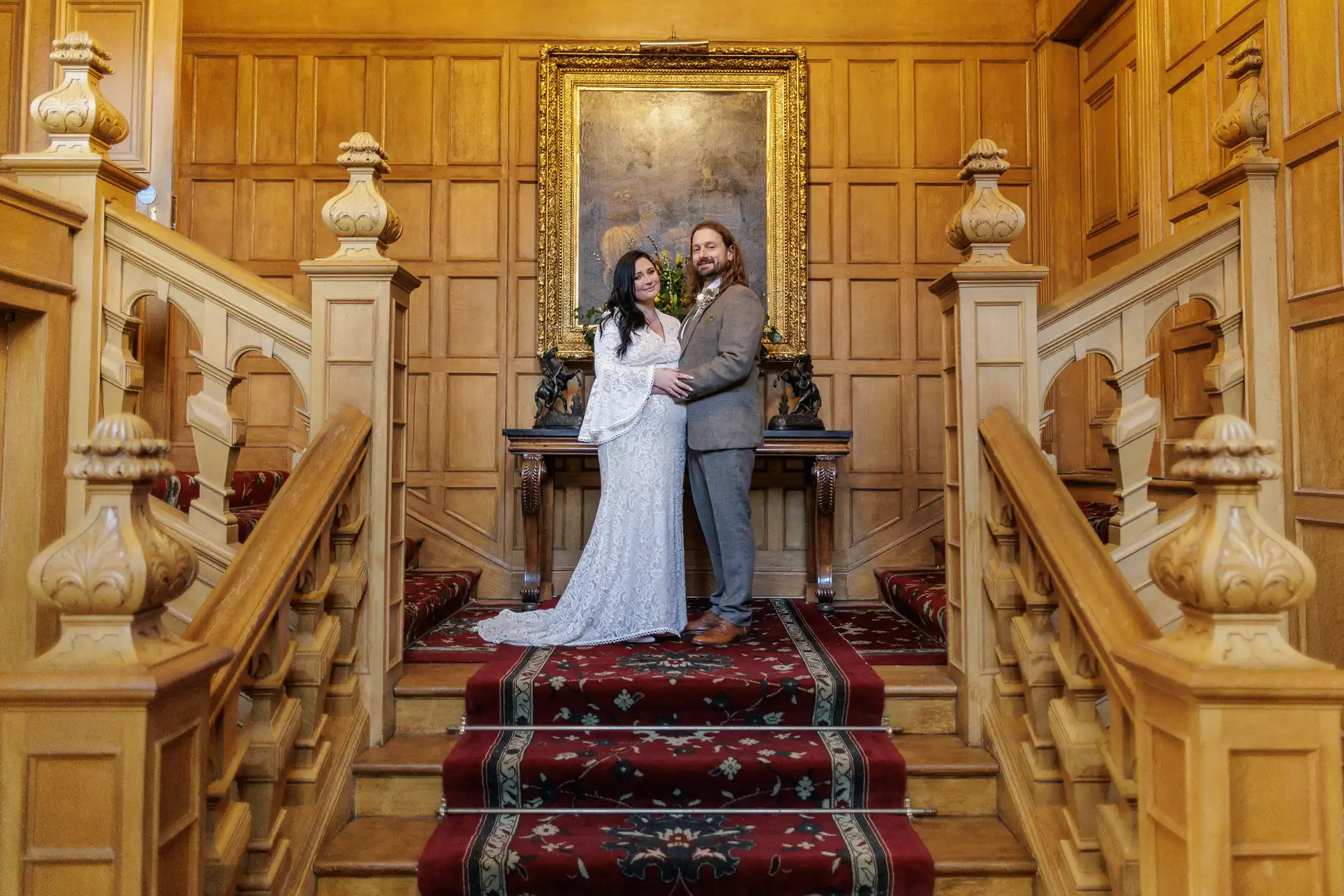 A couple in wedding attire stands at the centre of an ornate wooden staircase. The bride wears a white lace dress, and the groom is in a tan suit. Behind them is a large gold-framed painting on a wood-panelled wall, and the stairs are covered with a red patterned carpet.