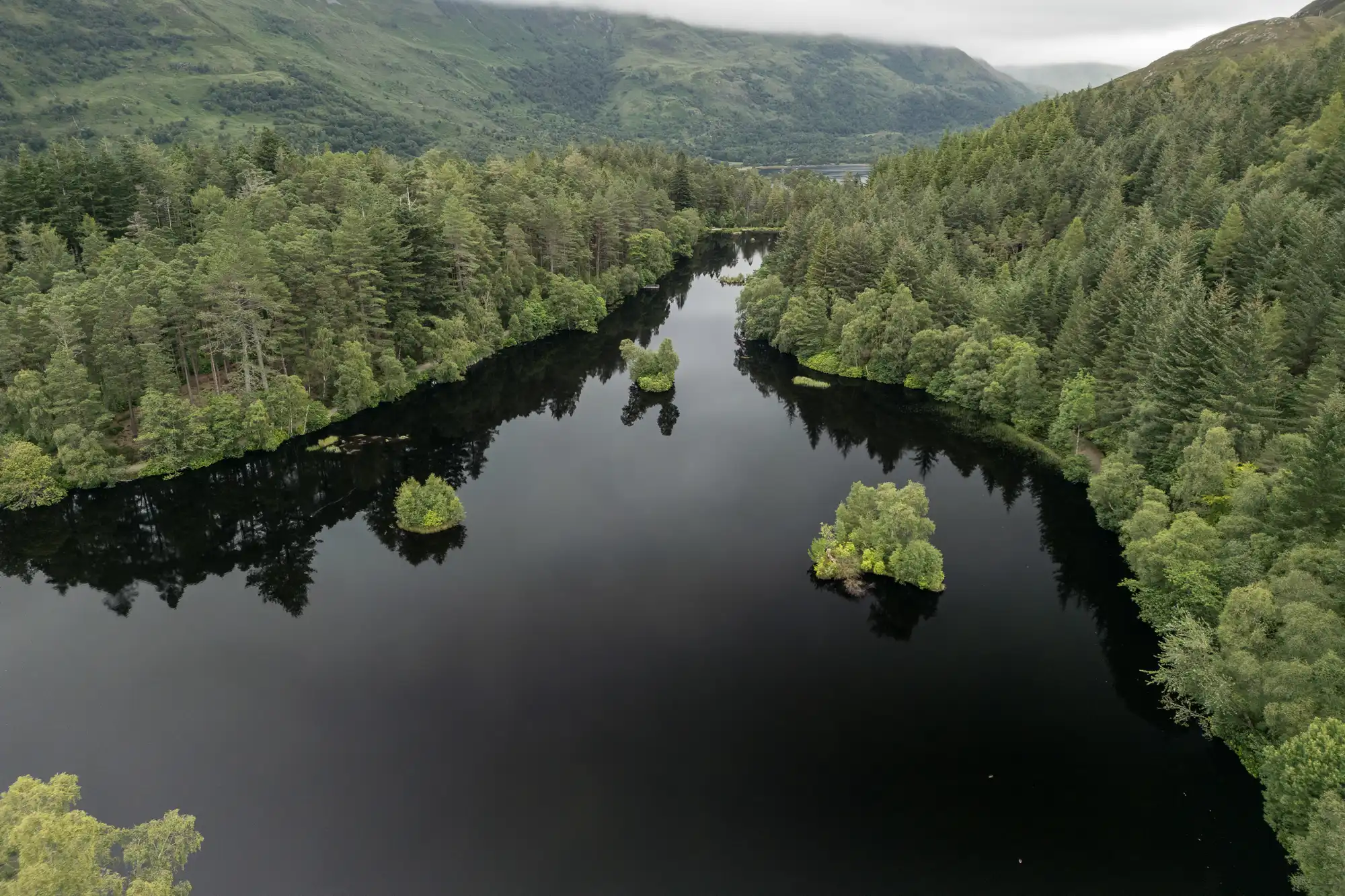 A calm, dark lake surrounded by dense green forest, with several small tree-covered islands in the water. Hills covered in trees and mist are visible in the background under a cloudy sky. The scene is tranquil and lush.