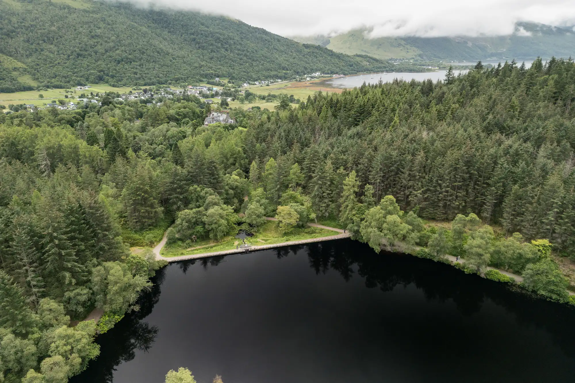 Aerial view of a calm, dark forest lake surrounded by dense green trees, with a winding path along its edge. In the background, a small village nestles at the base of misty, forested hills under a cloudy sky.