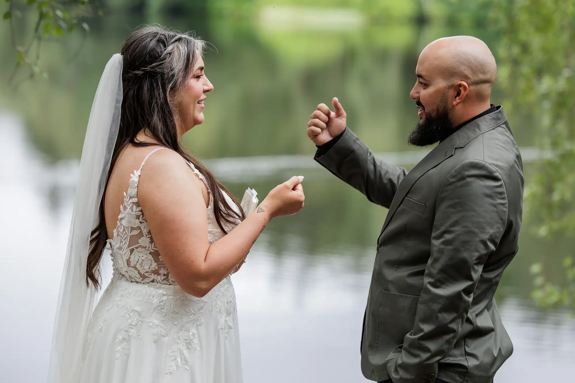 A bride in a white lace dress and veil faces a bald groom in a dark suit by a calm lake. Both are smiling and holding their hands as if about to play rock-paper-scissors, with green trees and water in the blurred background.