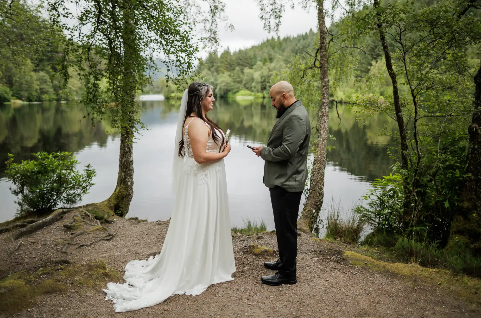 A bride in a white dress and a groom in a suit stand facing each other by a tranquil lakeside, surrounded by green trees and foliage. The groom reads from a piece of paper whilst the bride smiles, in a serene outdoor wedding setting.