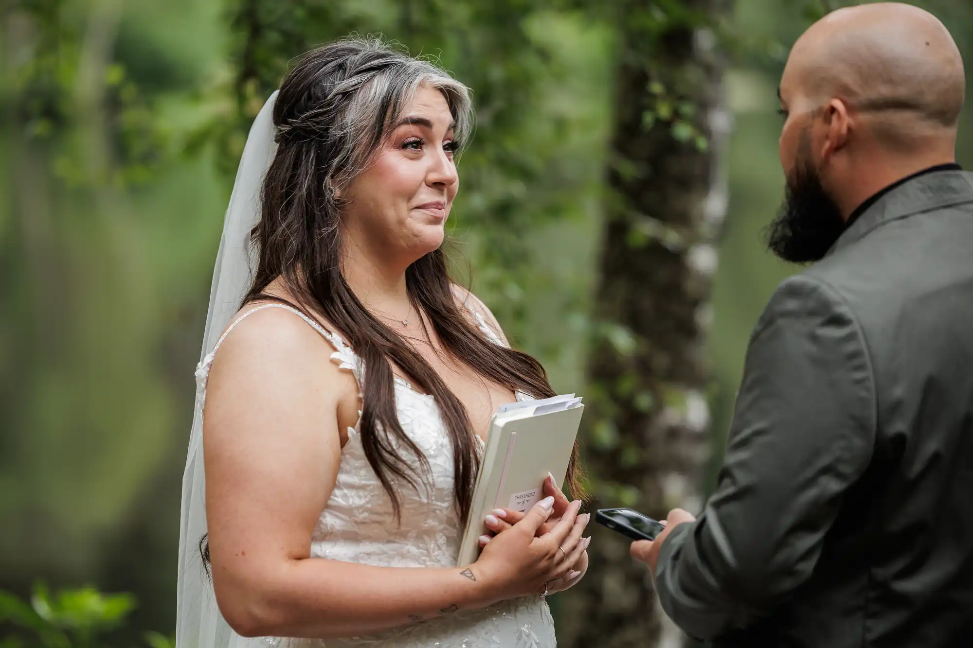 A bride in a white dress and veil holds a notebook, smiling emotionally at a groom with a beard, wearing a dark suit. They stand outdoors, surrounded by green trees, suggesting a heartfelt moment during their wedding ceremony.