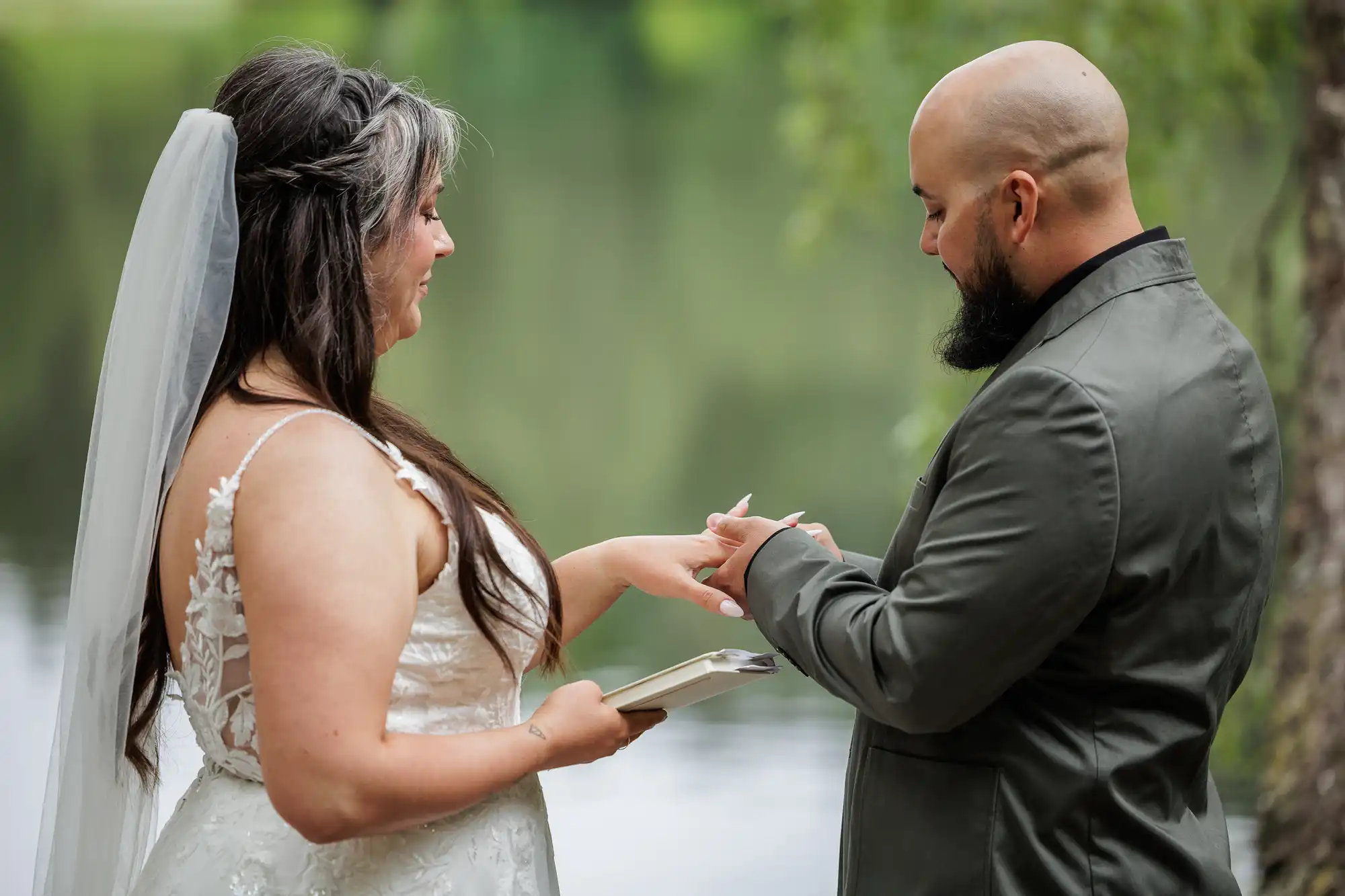 A bride in a white lace dress and veil holds a clipboard while a groom in a grey suit places a ring on her finger. They stand outdoors by a calm lake, surrounded by greenery, exchanging vows in a peaceful, intimate wedding moment.
