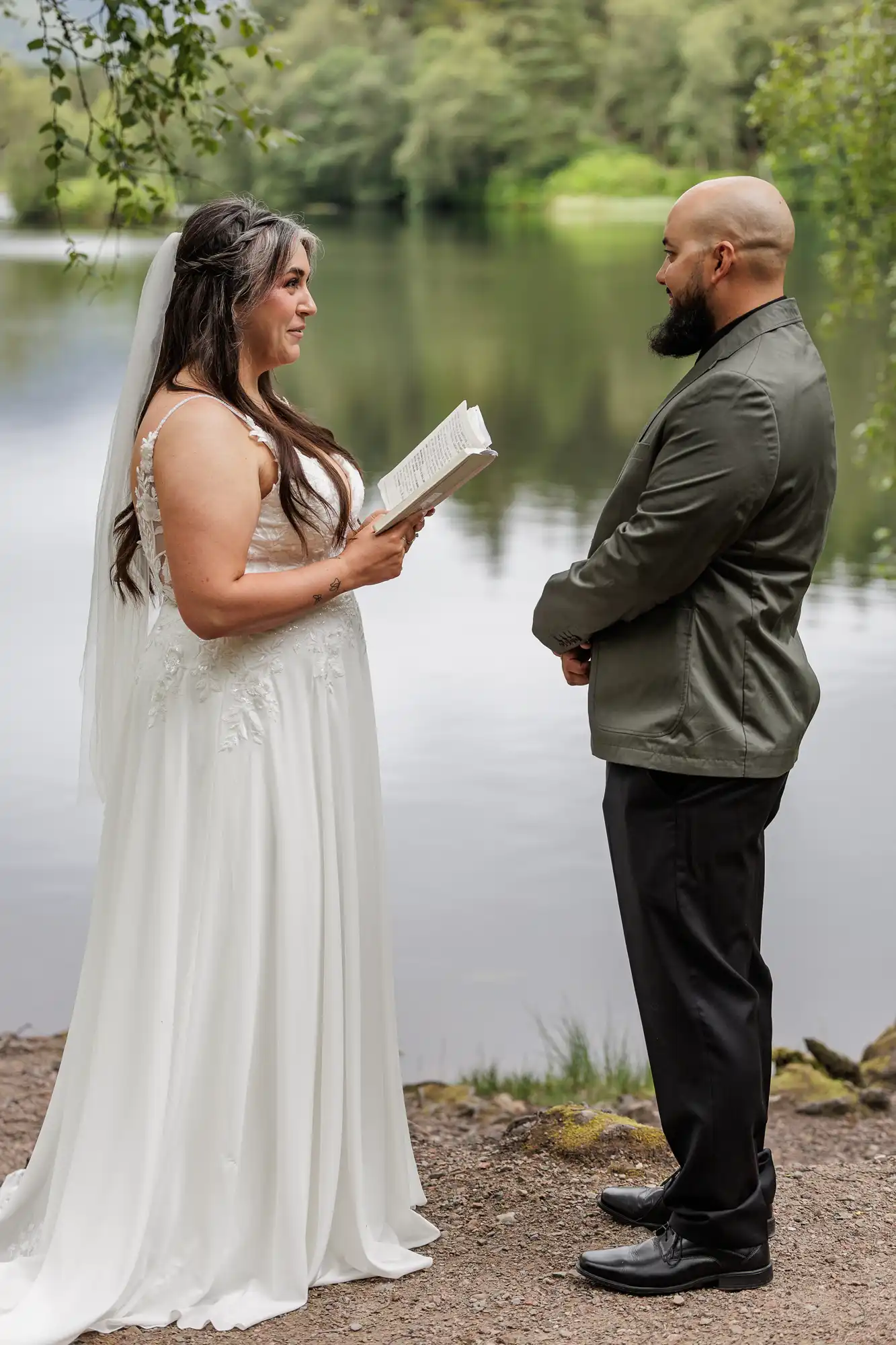 A bride in a white dress reads vows from a book to a groom in a green jacket and black trousers. They stand facing each other outdoors by a calm lake, with trees and greenery in the background on a cloudy day.