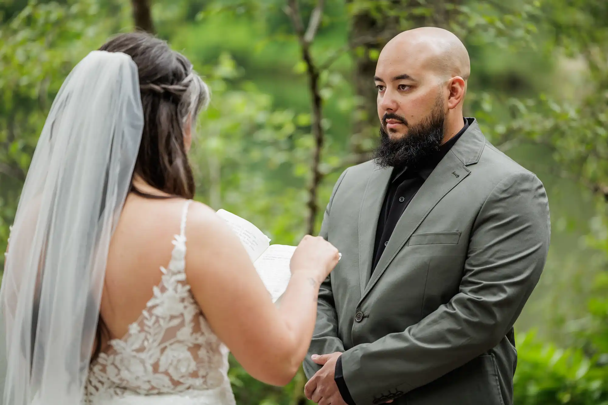 A bride in a white lace dress and veil reads vows from a notebook to a groom with a shaven head and beard, wearing a grey suit and black shirt. They stand outdoors surrounded by green trees, with the groom looking attentively at the bride.