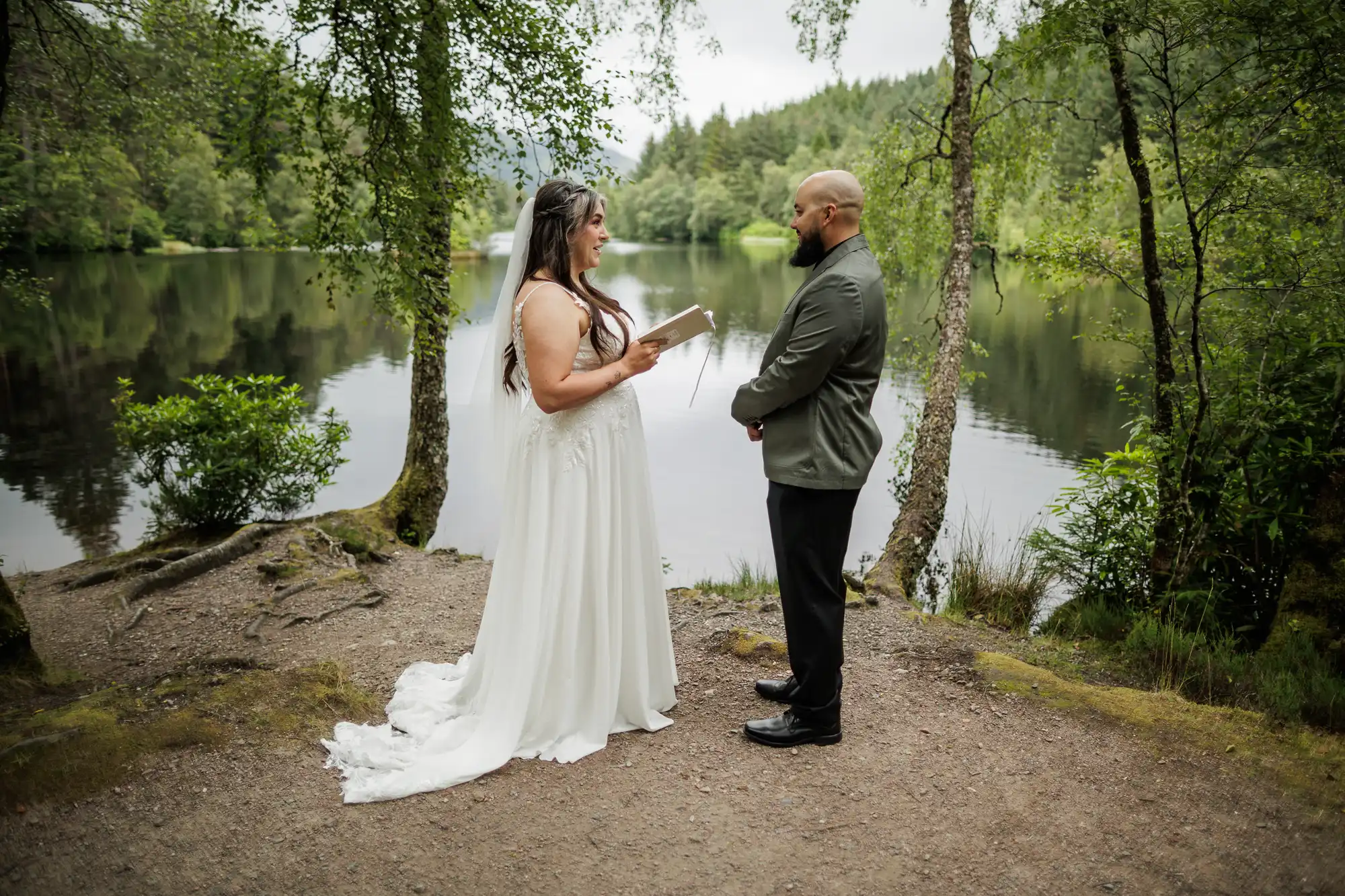 A bride in a white dress and veil reads vows to a groom in a dark suit. They stand facing each other on a forested lakeshore, surrounded by lush green trees with the calm water and distant woods in the background.