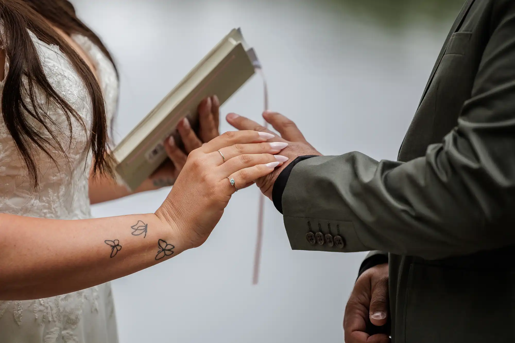 A bride in a white dress holds a book and gently touches the hand of a groom in a dark suit. Her arm shows butterfly tattoos and an engagement ring. The background is softly blurred, suggesting an outdoor setting near water.