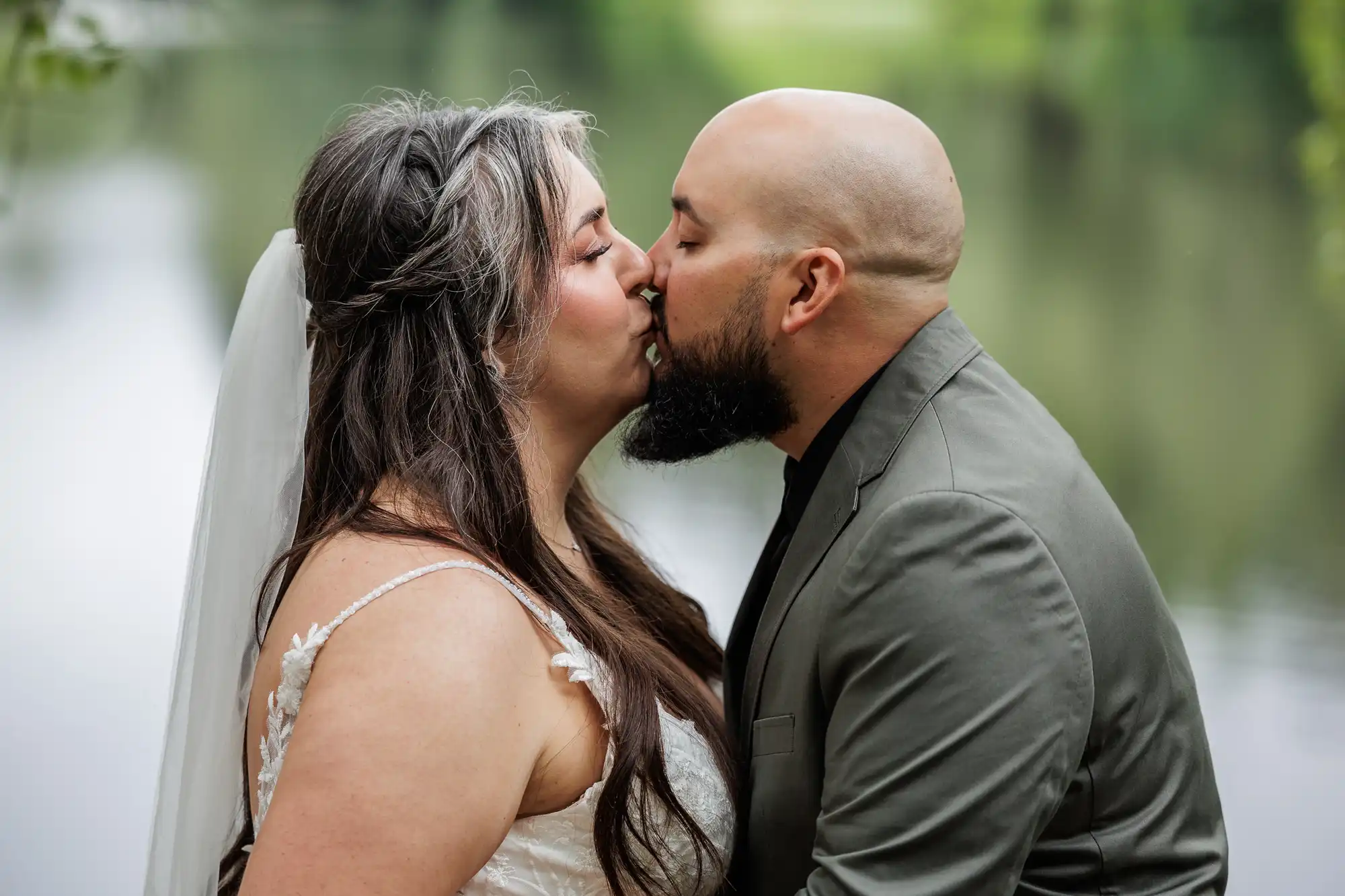 A bride in a white dress and veil and a groom in a grey suit kiss outdoors by a calm body of water, surrounded by soft greenery in the background. Their eyes are closed and they are embracing each other closely.