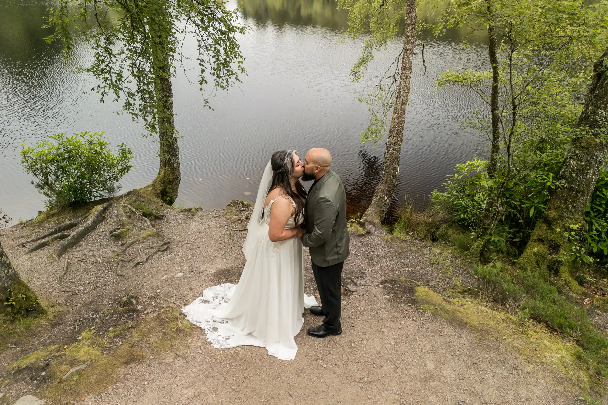 A bride in a white dress and veil kisses a groom in a suit on a dirt path by a calm, tree-lined lakeshore. Green foliage and tree roots surround them, and the water reflects the sky and trees, creating a serene, romantic scene.