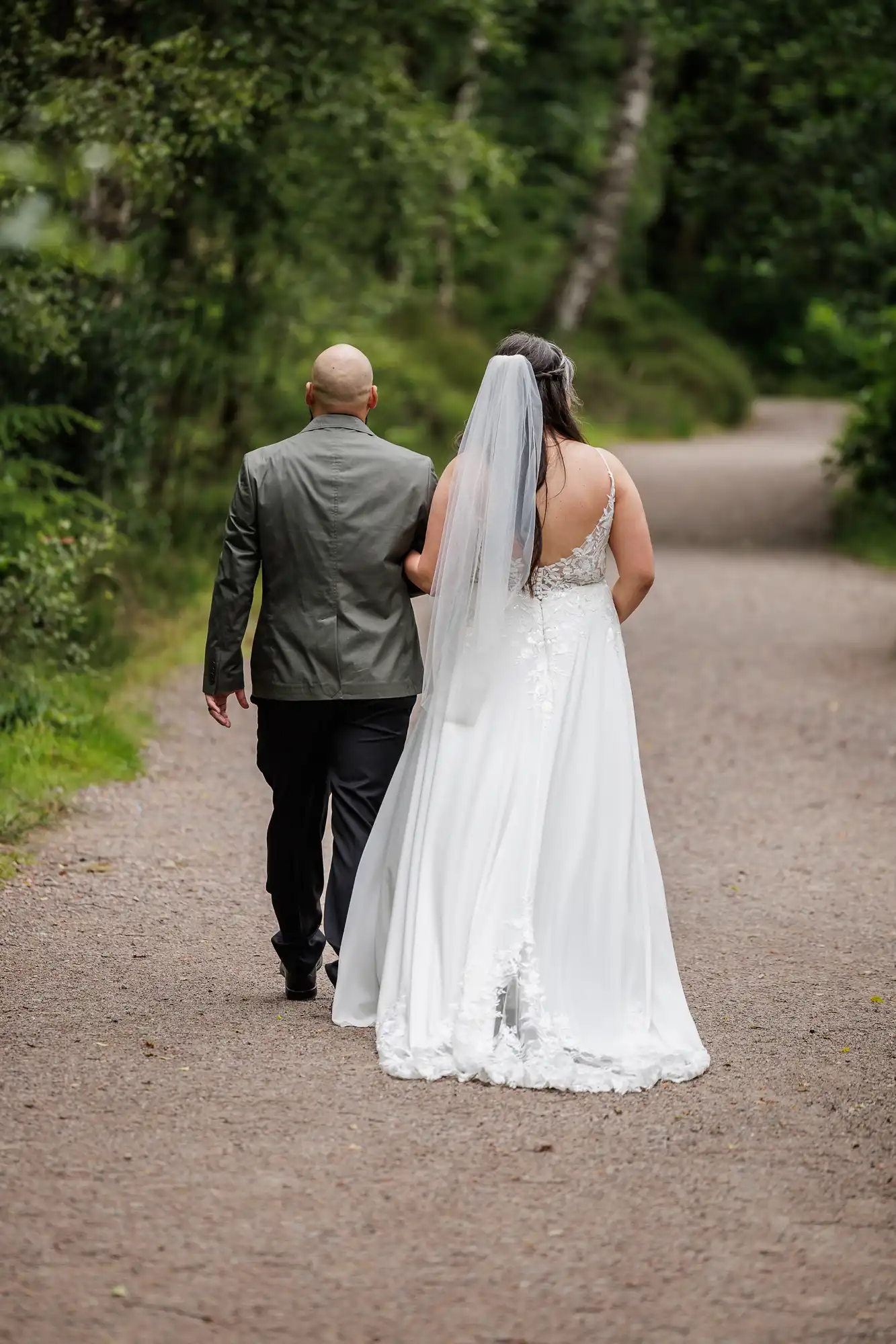 A bride in a white dress and veil walks arm-in-arm with a man in a dark suit along a tree-lined, gravel path, both seen from behind in a lush, green outdoor setting.