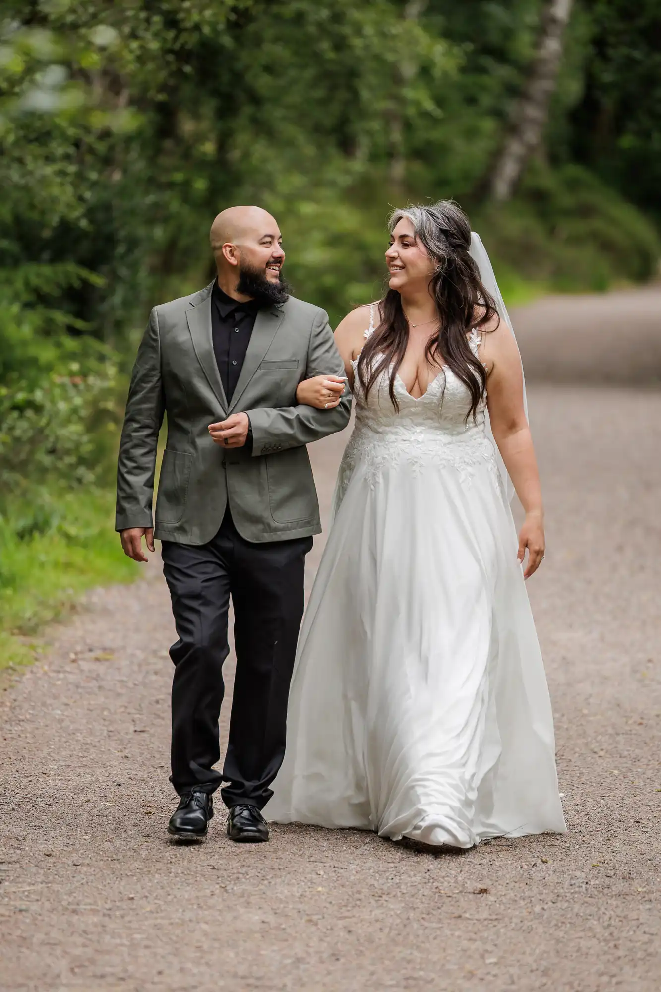 A smiling bride in a white gown and veil walks arm-in-arm with a bearded man in a green jacket and black trousers on a tree-lined path. Both look at each other happily. The background is lush and green, suggesting a peaceful outdoor setting.