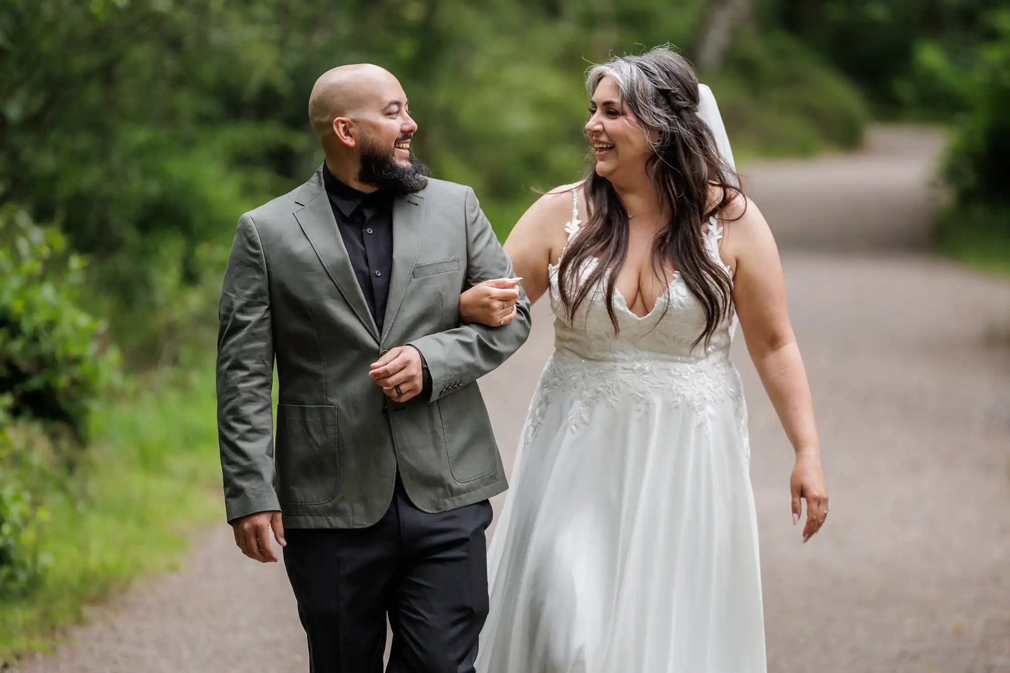 A smiling bride in a white lace wedding dress and veil walks arm-in-arm with a bearded groom in a green suit jacket and black shirt on a tree-lined path, both looking at each other happily.