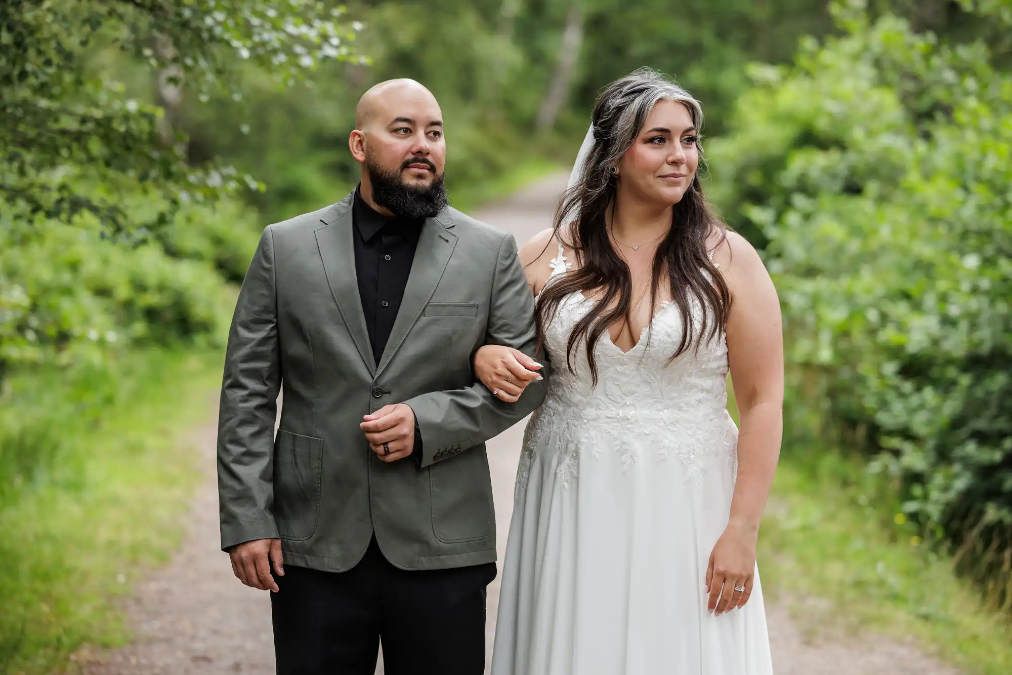 A bride in a white lace wedding dress with a veil stands arm-in-arm with a groom in a grey suit and black shirt. They are outdoors on a wooded path, surrounded by lush green foliage, both looking thoughtfully into the distance.