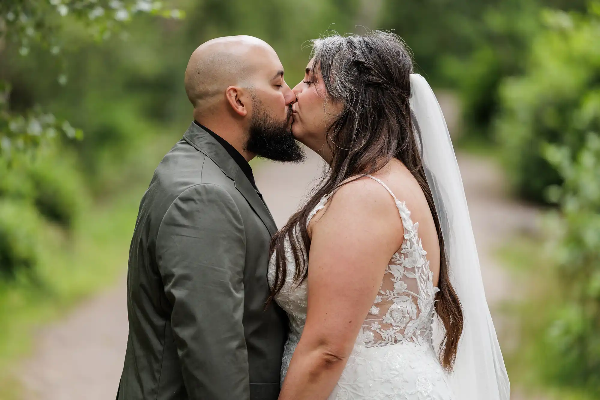 A couple snogs outdoors on a wooded path. The person on the left is bald with a beard and wears a grey suit. The person on the right has long dark hair with grey streaks, a white lace wedding dress, and a long veil. Lush greenery surrounds them.
